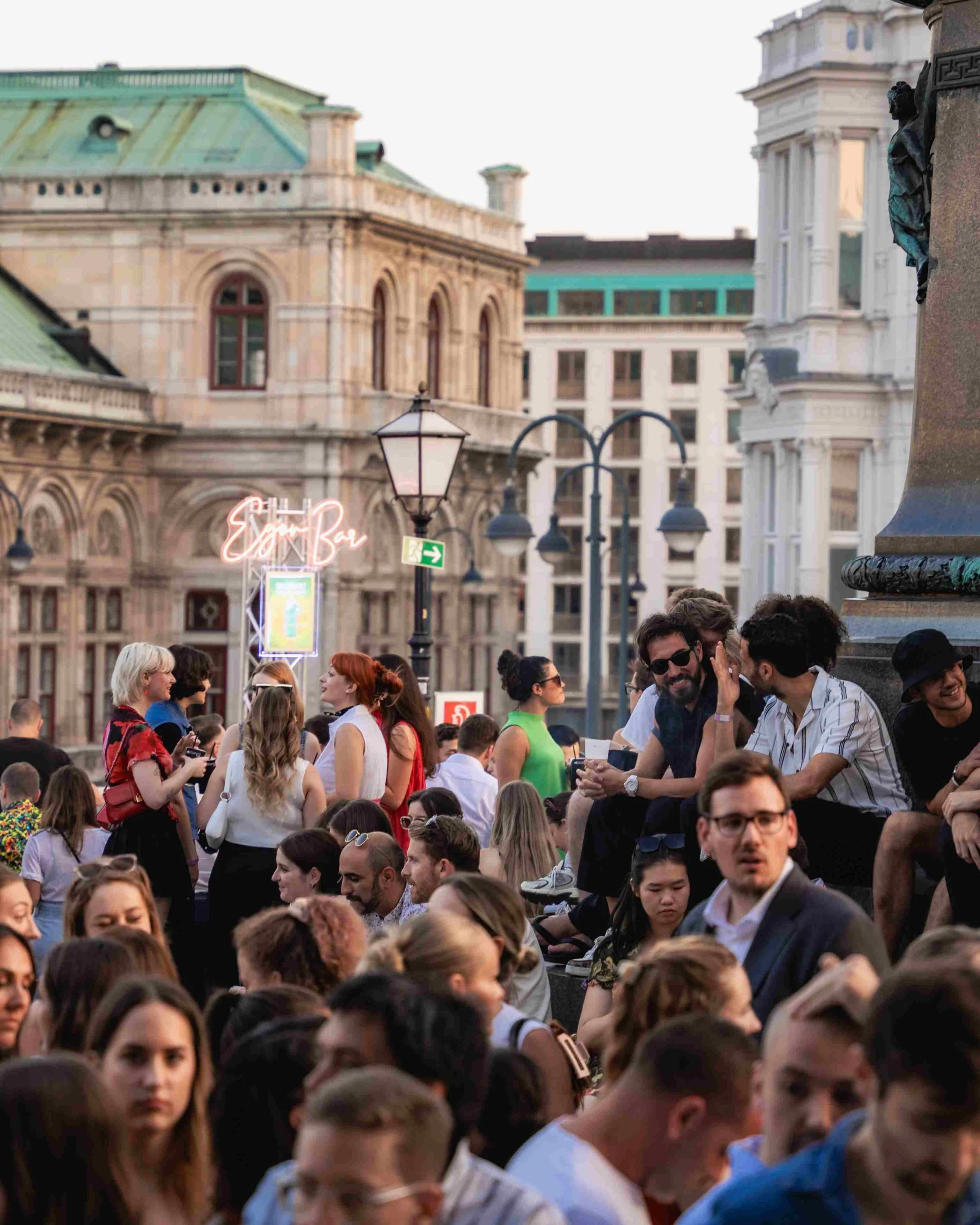 A crowd of people gathered outdoors in front of historic buildings with detailed architecture. Some individuals are engaged in conversation, while others look around or use their phones. The scene appears lively and social, with the background showin