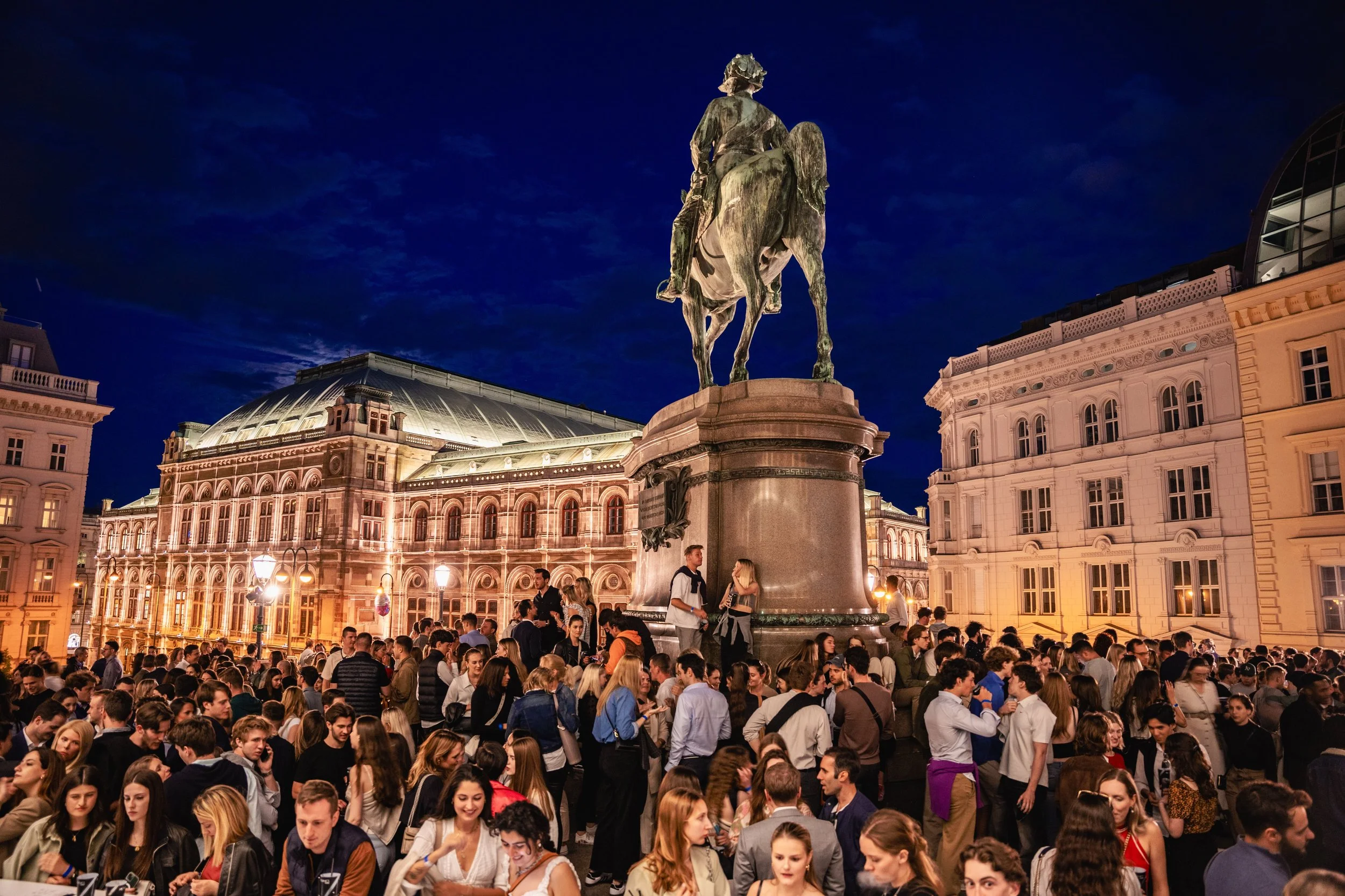 A large crowd of people gathered around an equestrian statue at night in an urban area with historical architecture and a clear sky.
