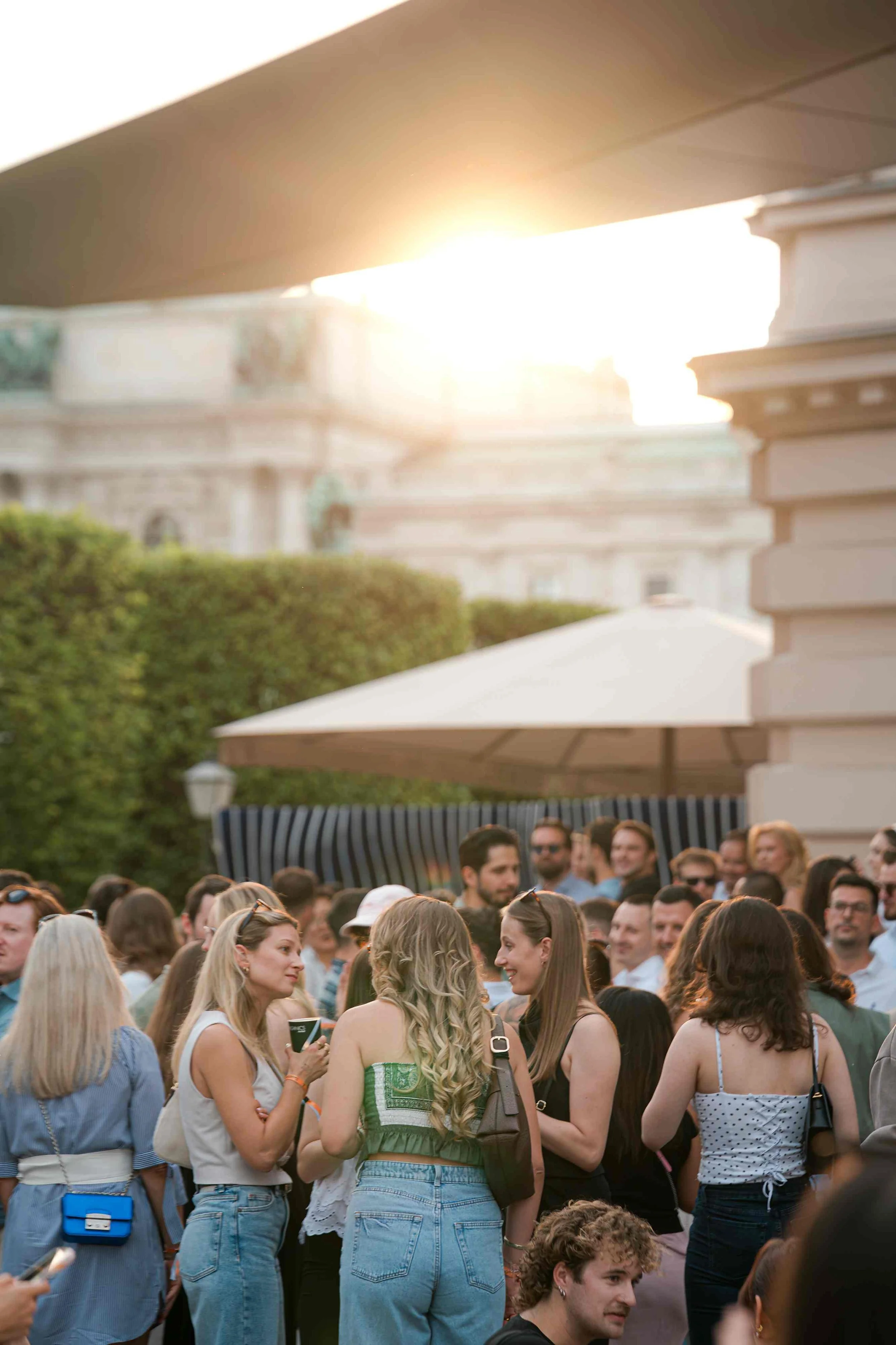 A large group of people gathered outdoors on a warm sunny day, with an umbrella and greenery visible in the background.