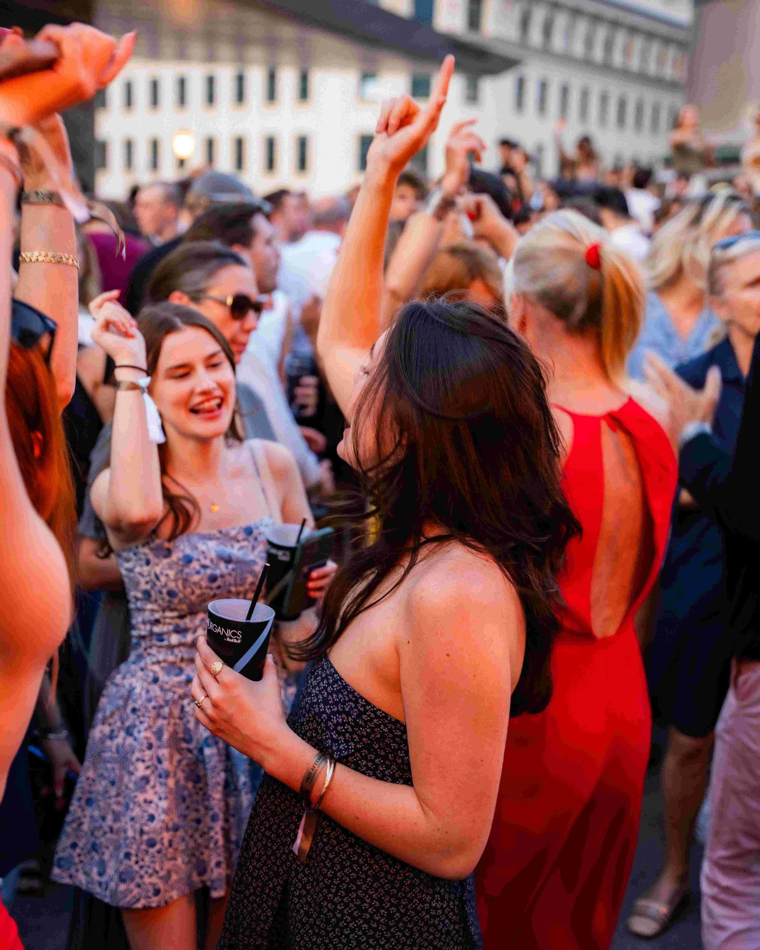 A crowd of people attending an outdoor event in the evening, with some raising their hands and smiling, dressed in casual summer attire.