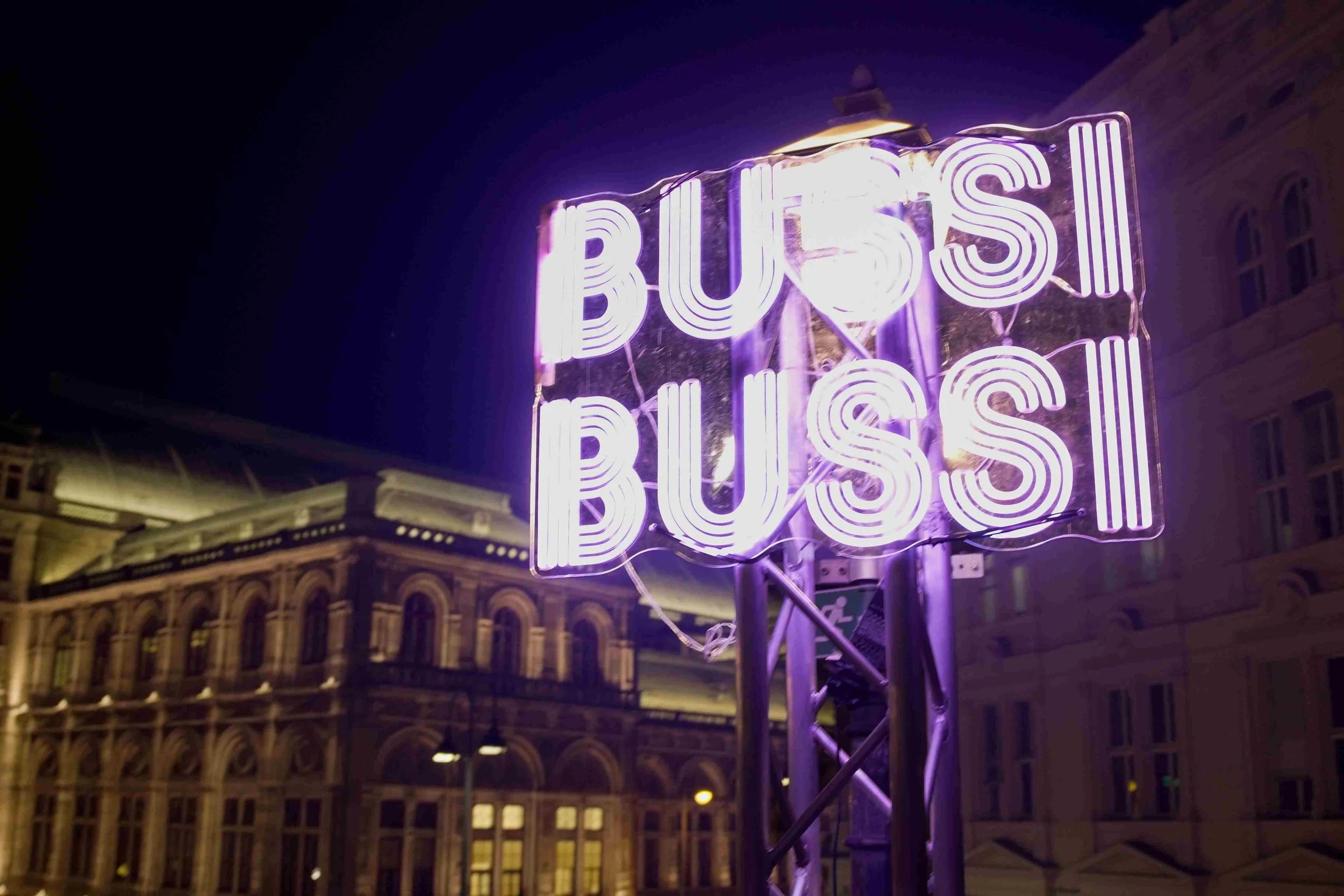 A neon sign that reads 'BISS' in purple lights, mounted on a metal frame outdoors at night, with historic buildings in the background.