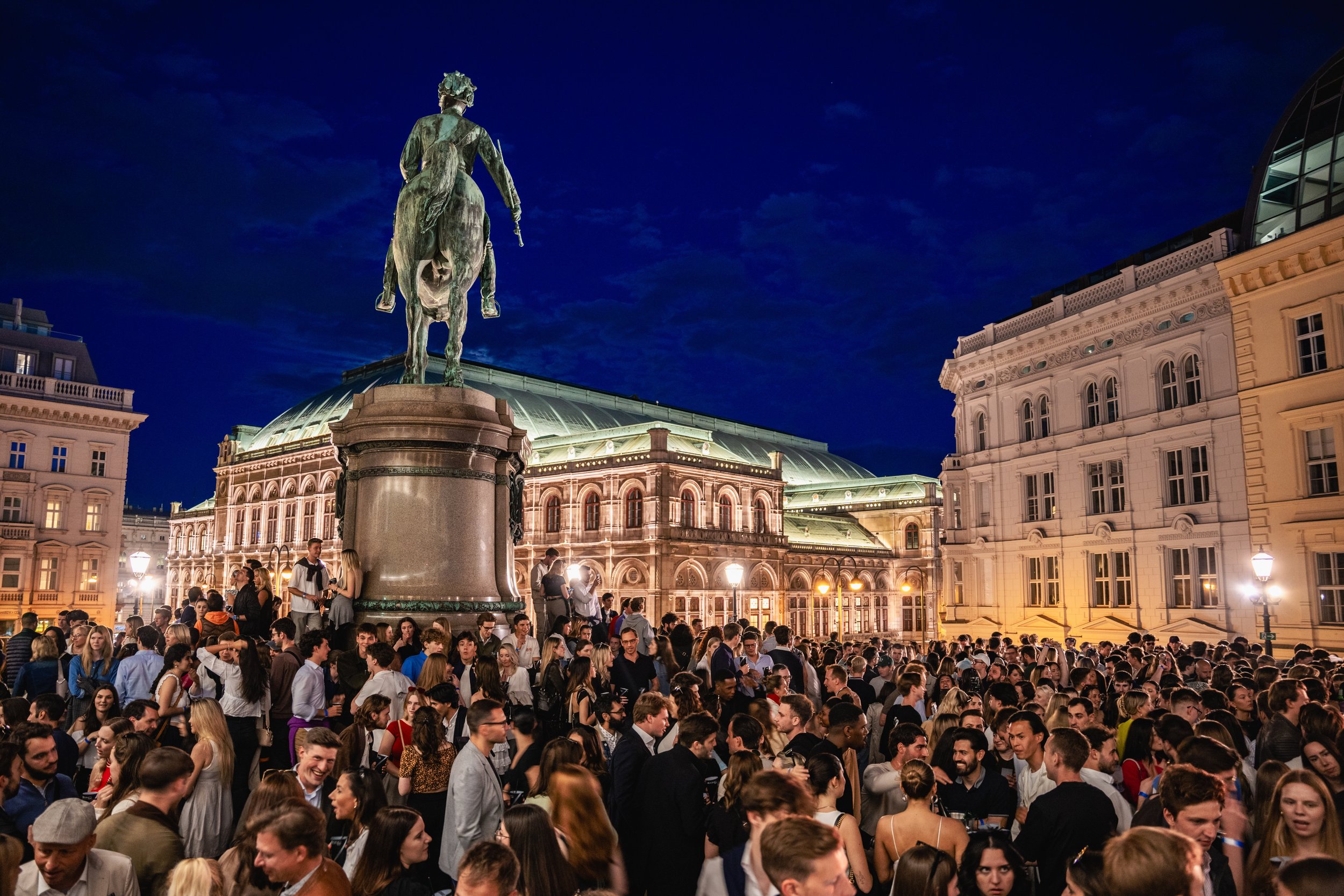 A large crowd of people gathered in a city square at night, with a statue of a man on a horse in the center. The surrounding buildings are historic and illuminated, and the sky is dark with some clouds.