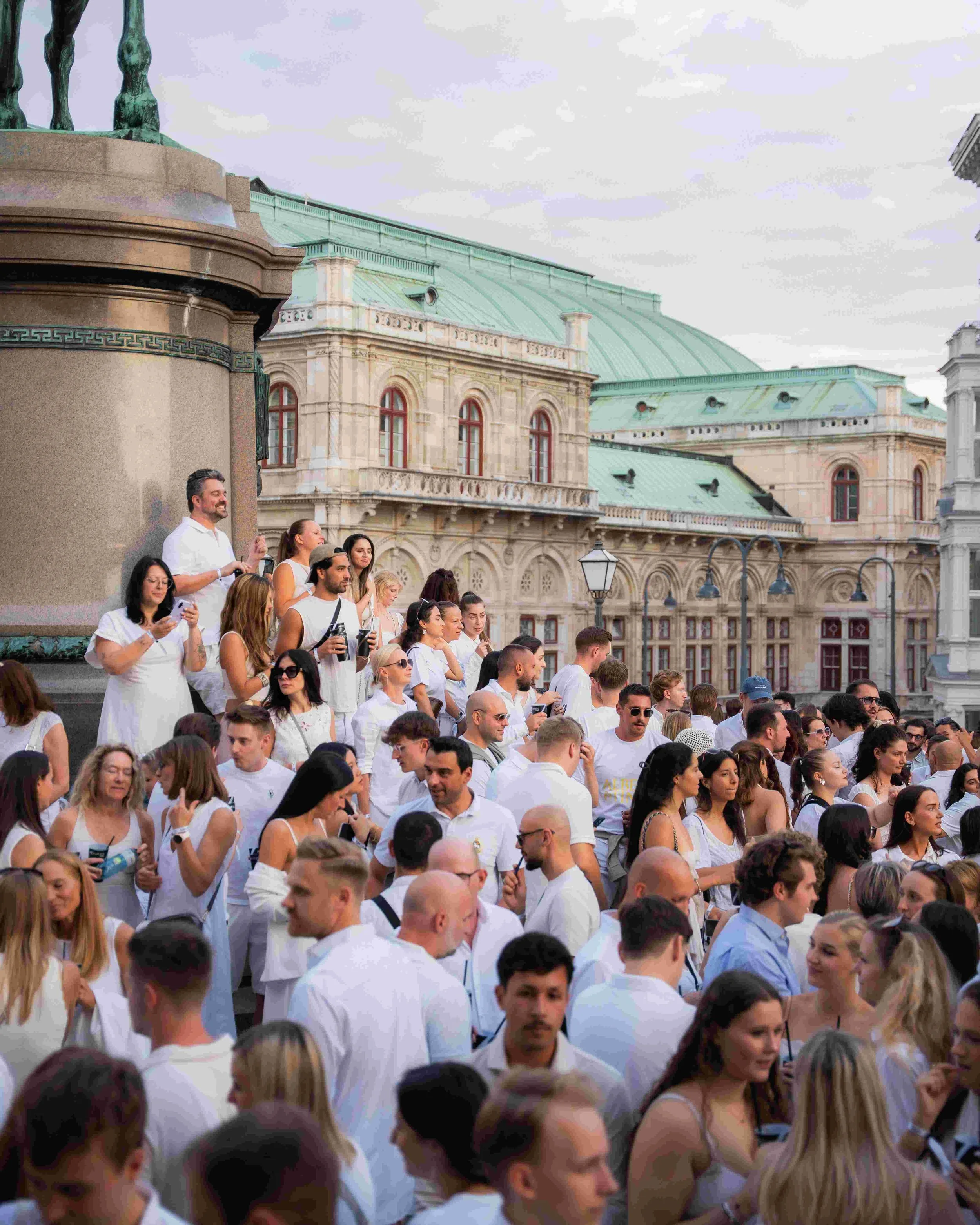 A crowd of people gathered outdoors in front of a historic building with a green roof and large windows. Some people are wearing white shirts, and many are holding drinks or taking selfies. The background features cloudy skies and part of a statue.
