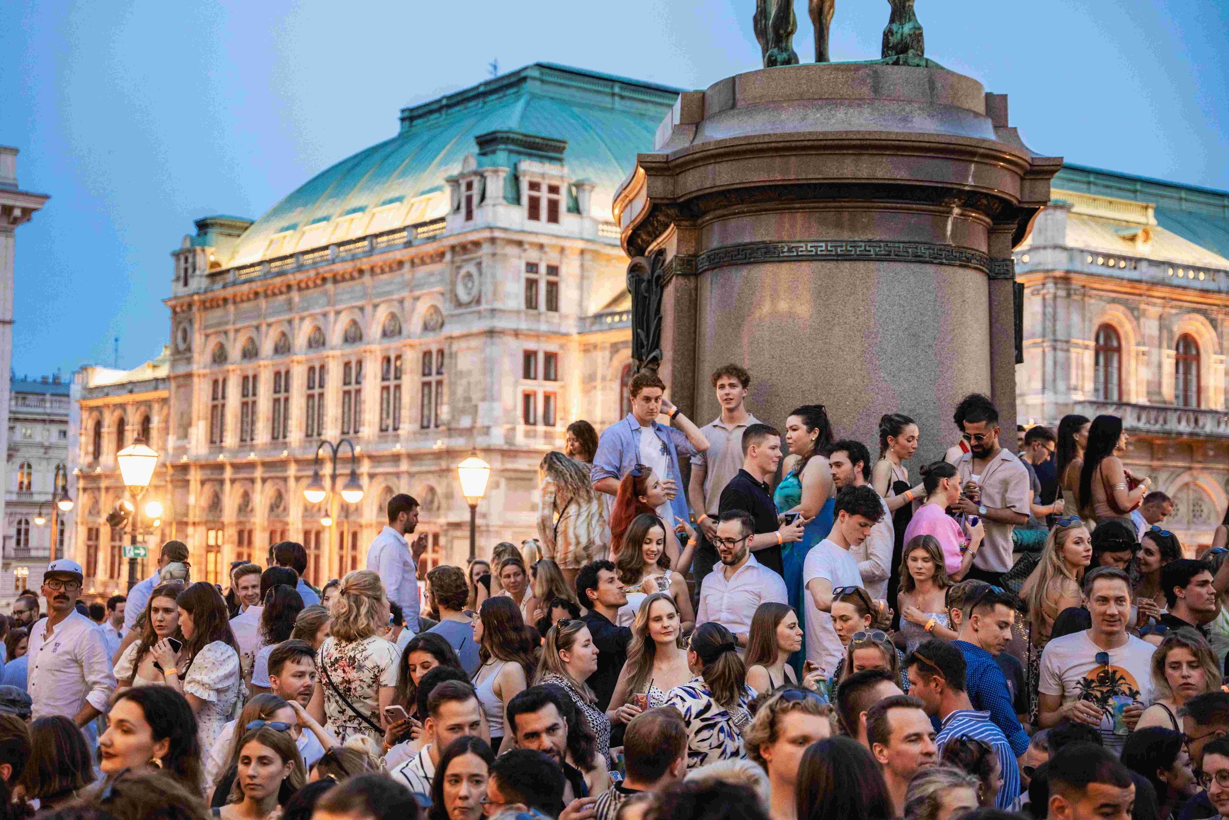 A large crowd of diverse people gathered outdoors in front of a historic building with a greenish dome and ornate architecture during the evening.