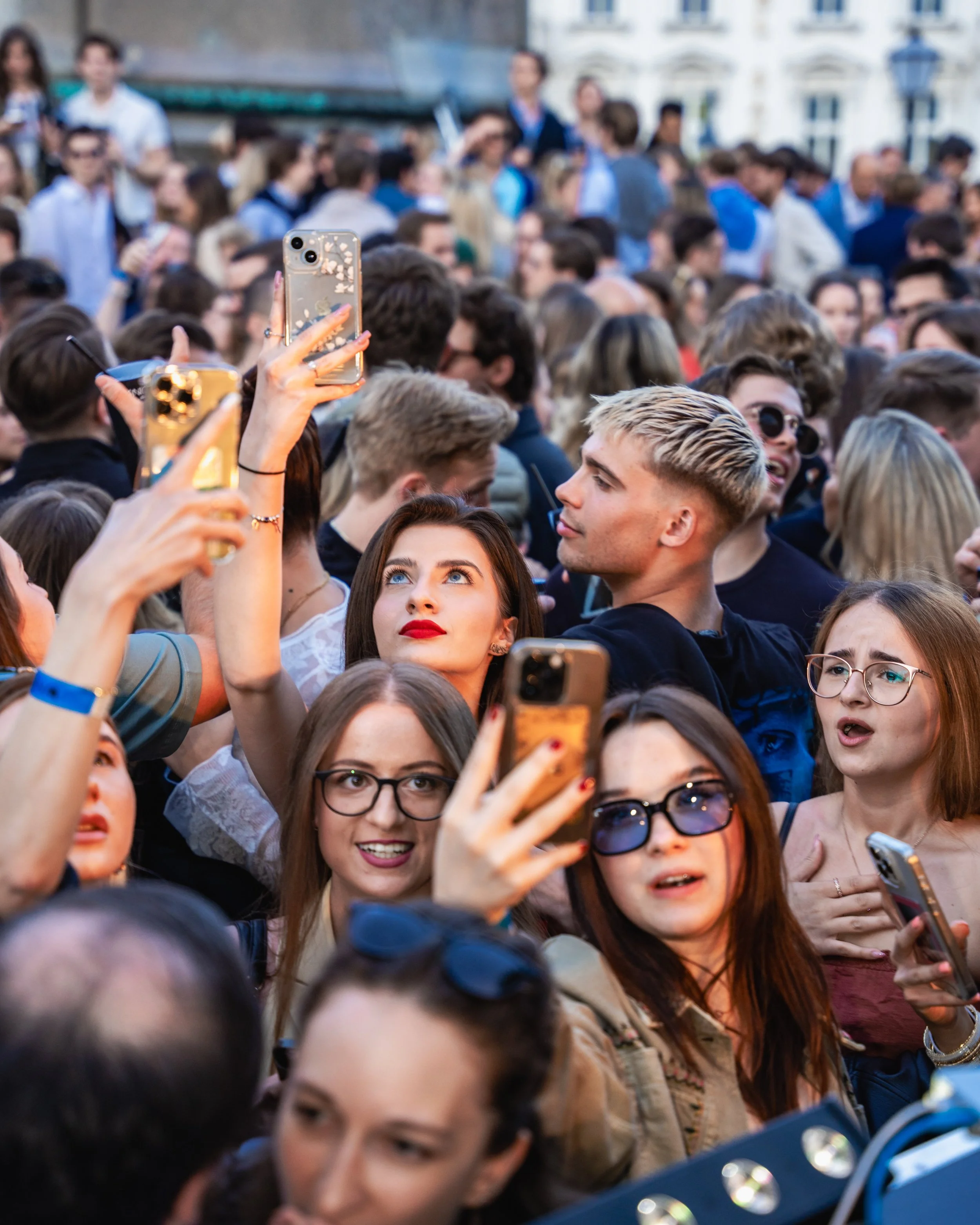 A crowd of people gathered outdoors, many taking photos or videos with their smartphones, and some wearing sunglasses and casual clothing.