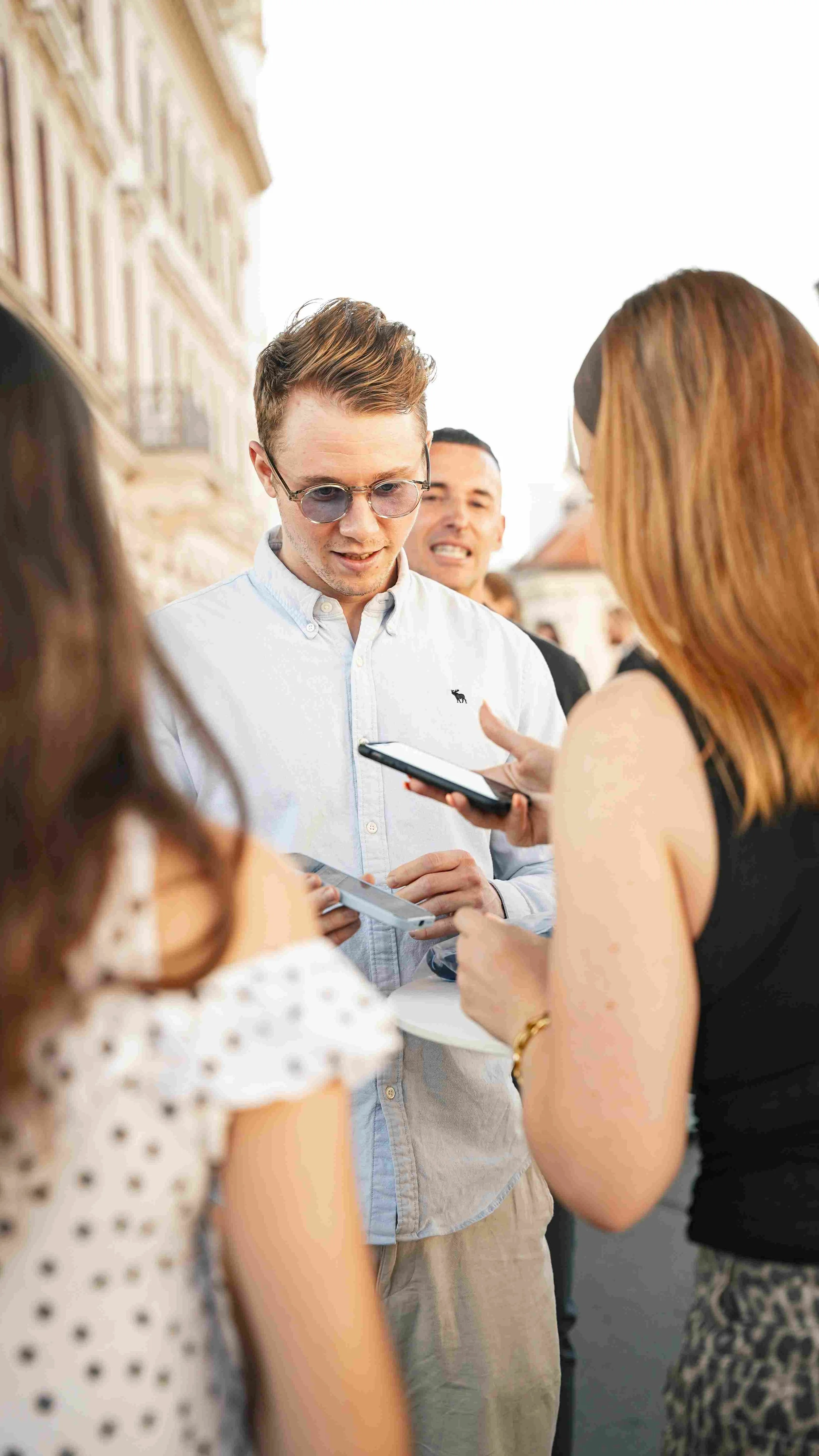 A group of people gathered outdoors, with a man in a white shirt and sunglasses speaking to a woman with reddish hair, as others listen near historic buildings.