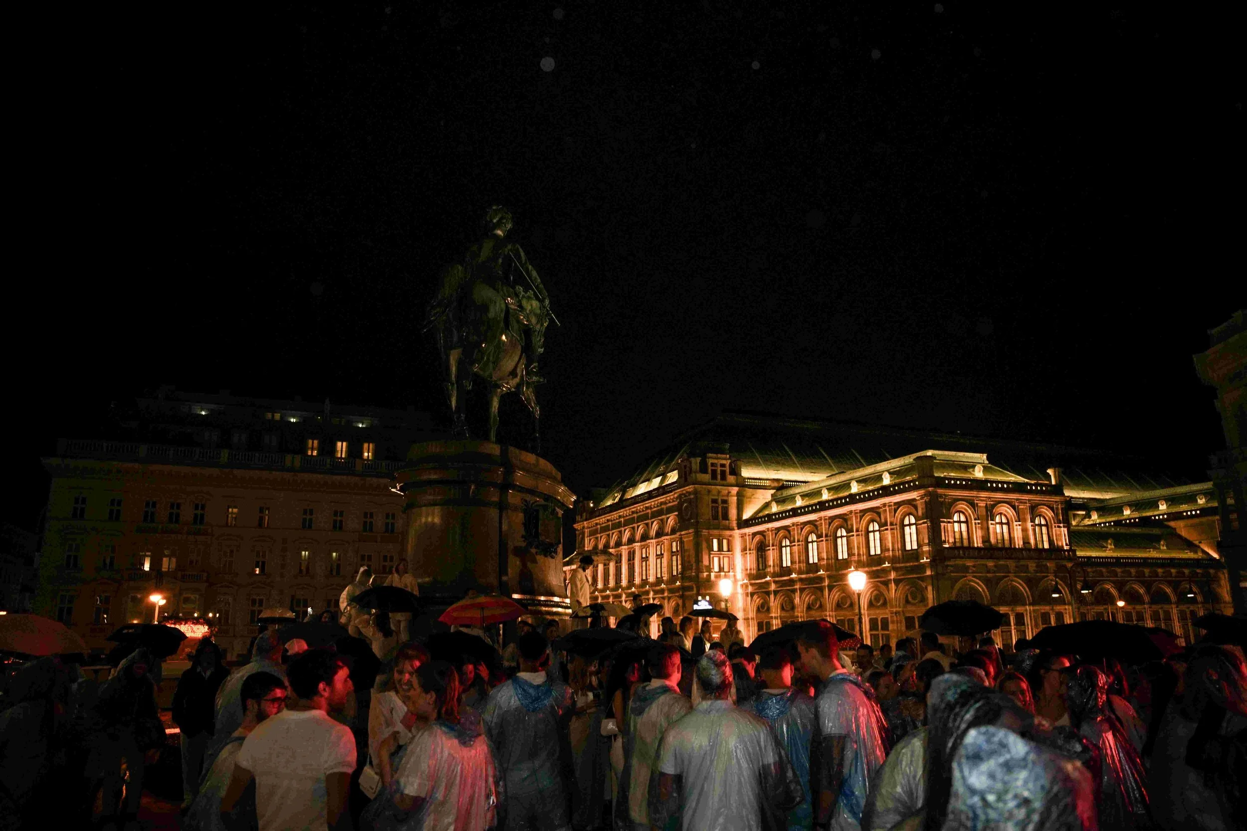 Nighttime scene in a city square with a crowd of people under umbrellas, illuminated historic buildings, and a statue on a pedestal.