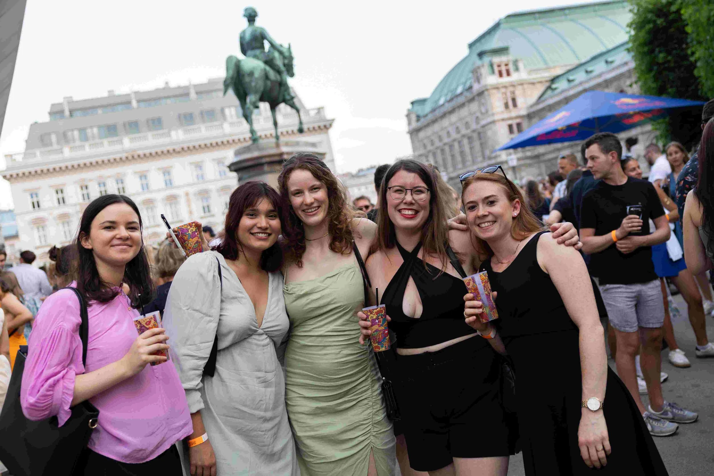 Group of five young women smiling and posing for a photo at an outdoor event with a historic building and a statue of a man on a horse in the background. Some women hold colorful cups, and others have sunglasses or bags.