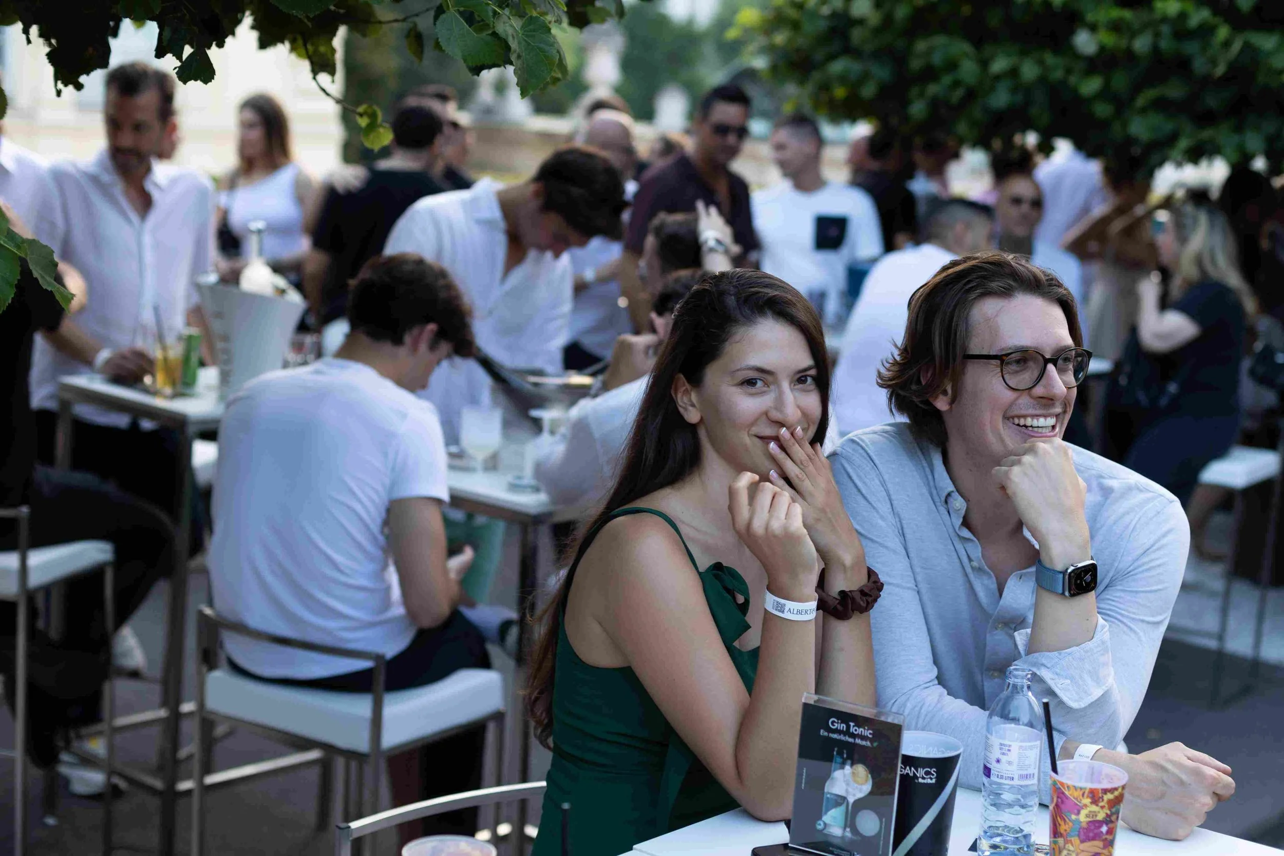 People gathered at an outdoor event, sitting at tables under trees, drinking beverages, and smiling, with some engaging in conversation.