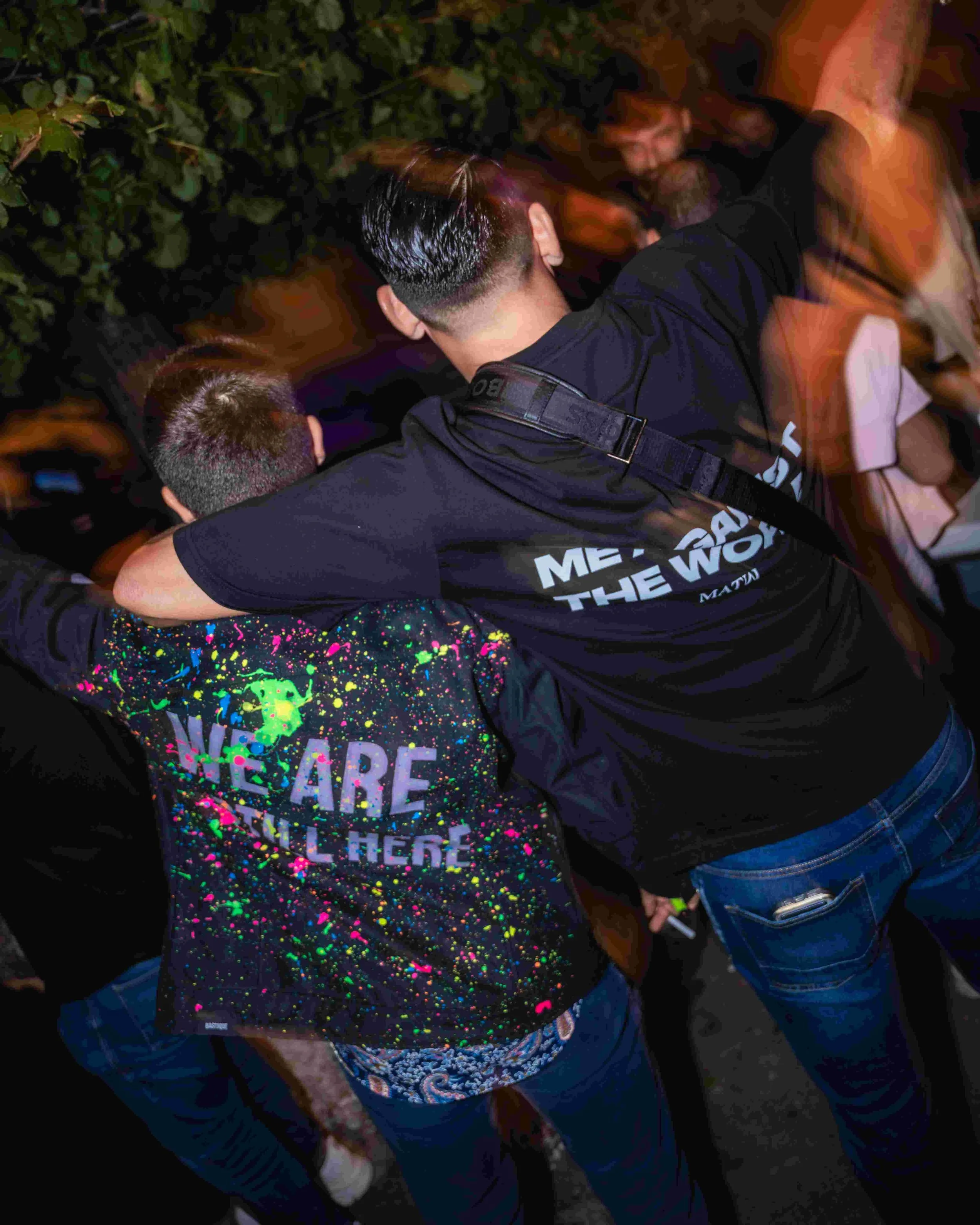 People hugging outdoors at night, one person with black hair and another with short blonde hair, wearing casual clothes, surrounded by greenery.