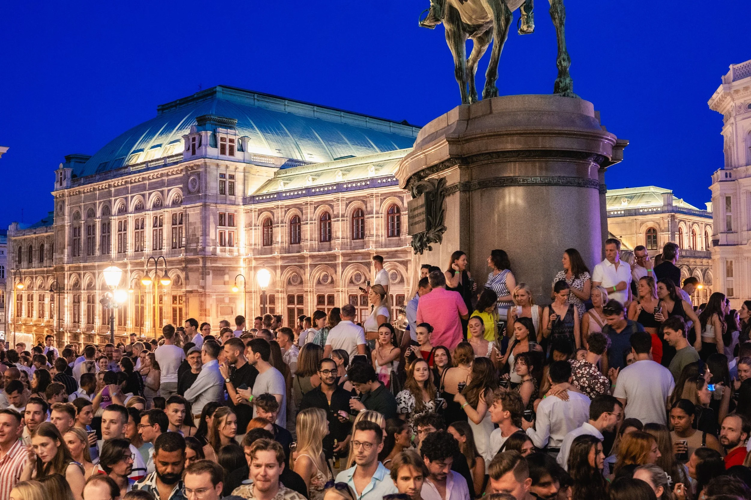 A large crowd of people gathered outdoors during the evening, with historic architecture and an illuminated building with a domed roof in the background.