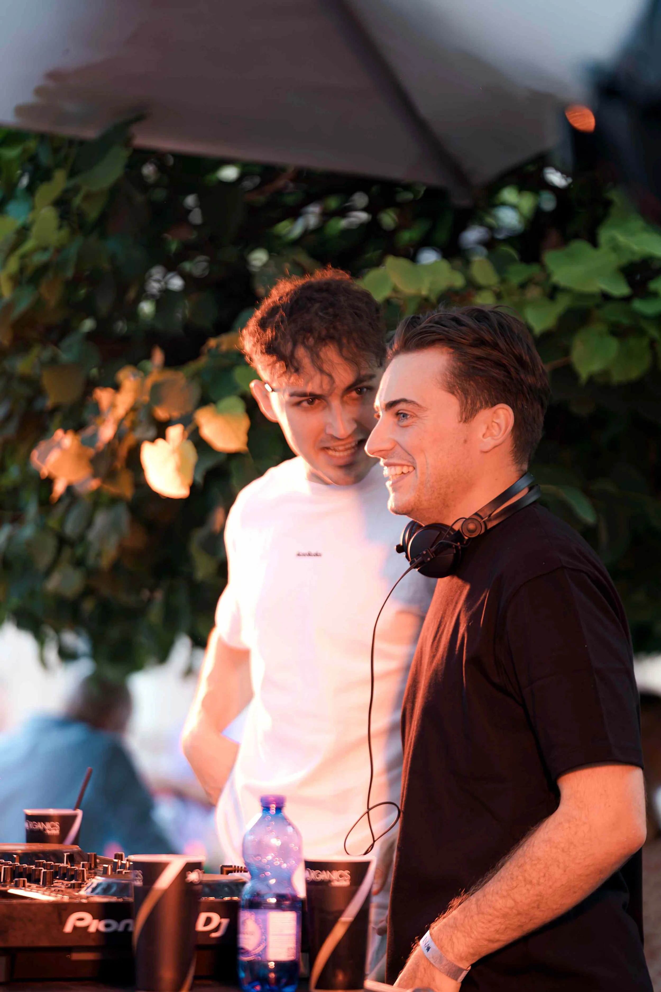 Two young men at a DJ booth outdoors, with one wearing headphones around his neck, scene lit by natural sunset light, green foliage in background.