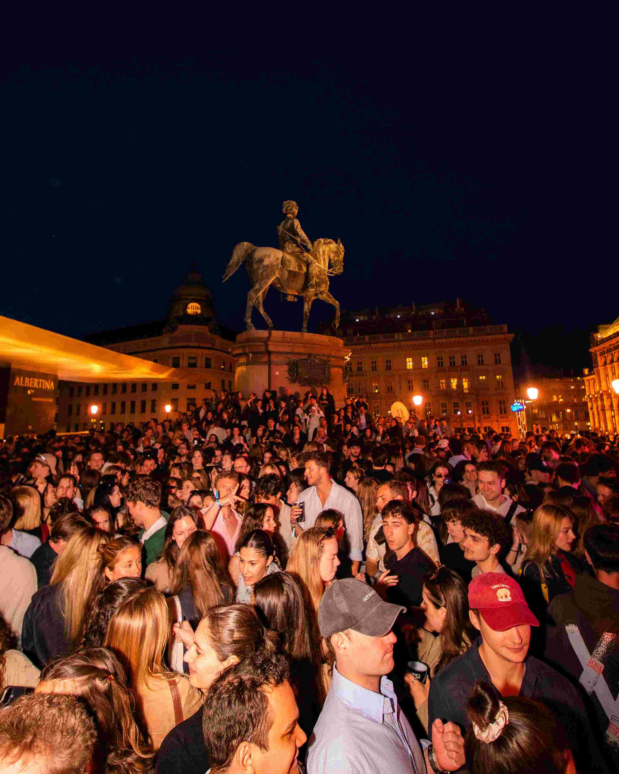 Crowd of people gathered outdoors at night in front of an equestrian statue at a public event or celebration, with buildings illuminated in the background.