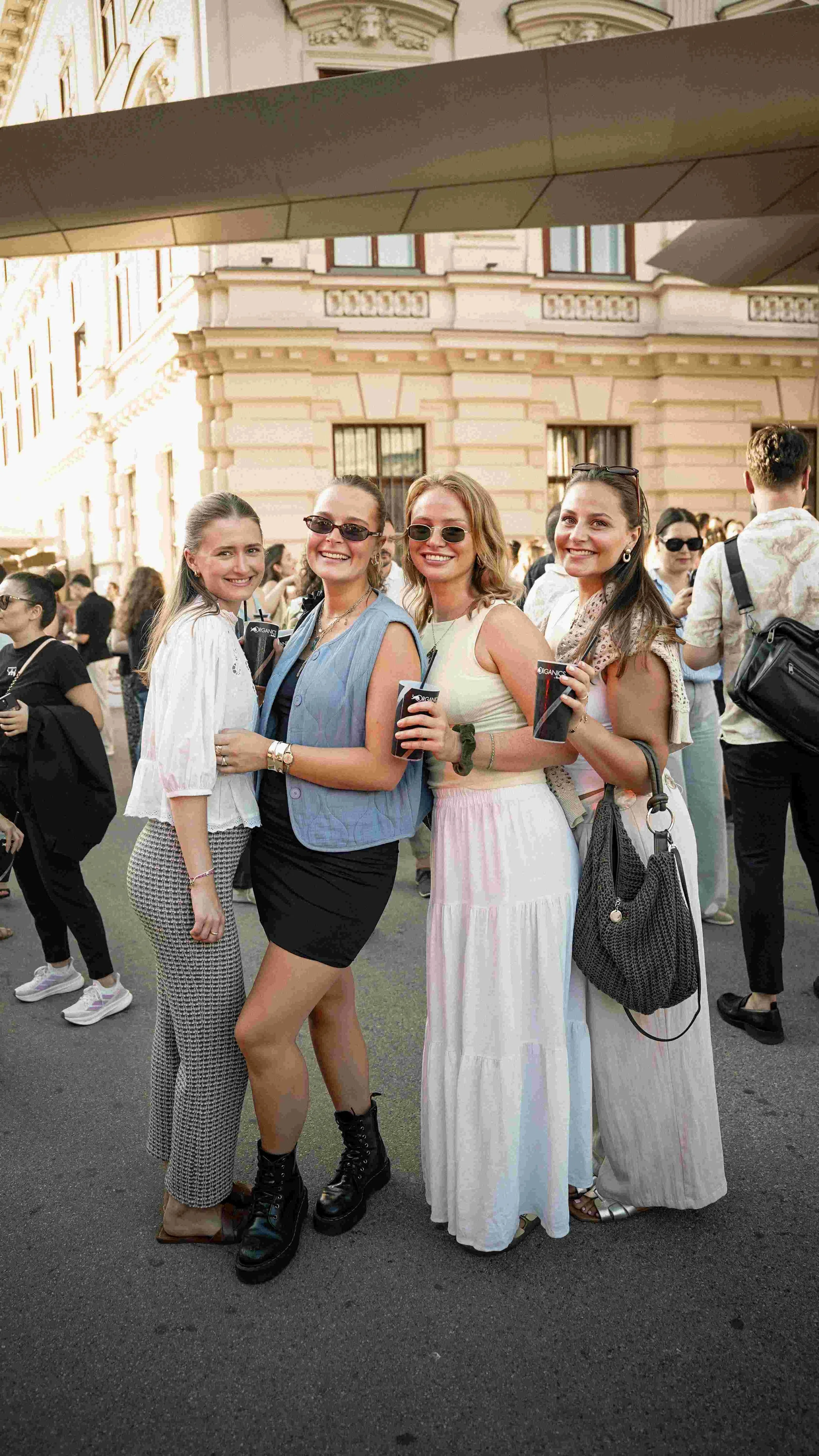 Four women standing together at an outdoor event, smiling and holding drinks, with a crowd and historic building behind them.