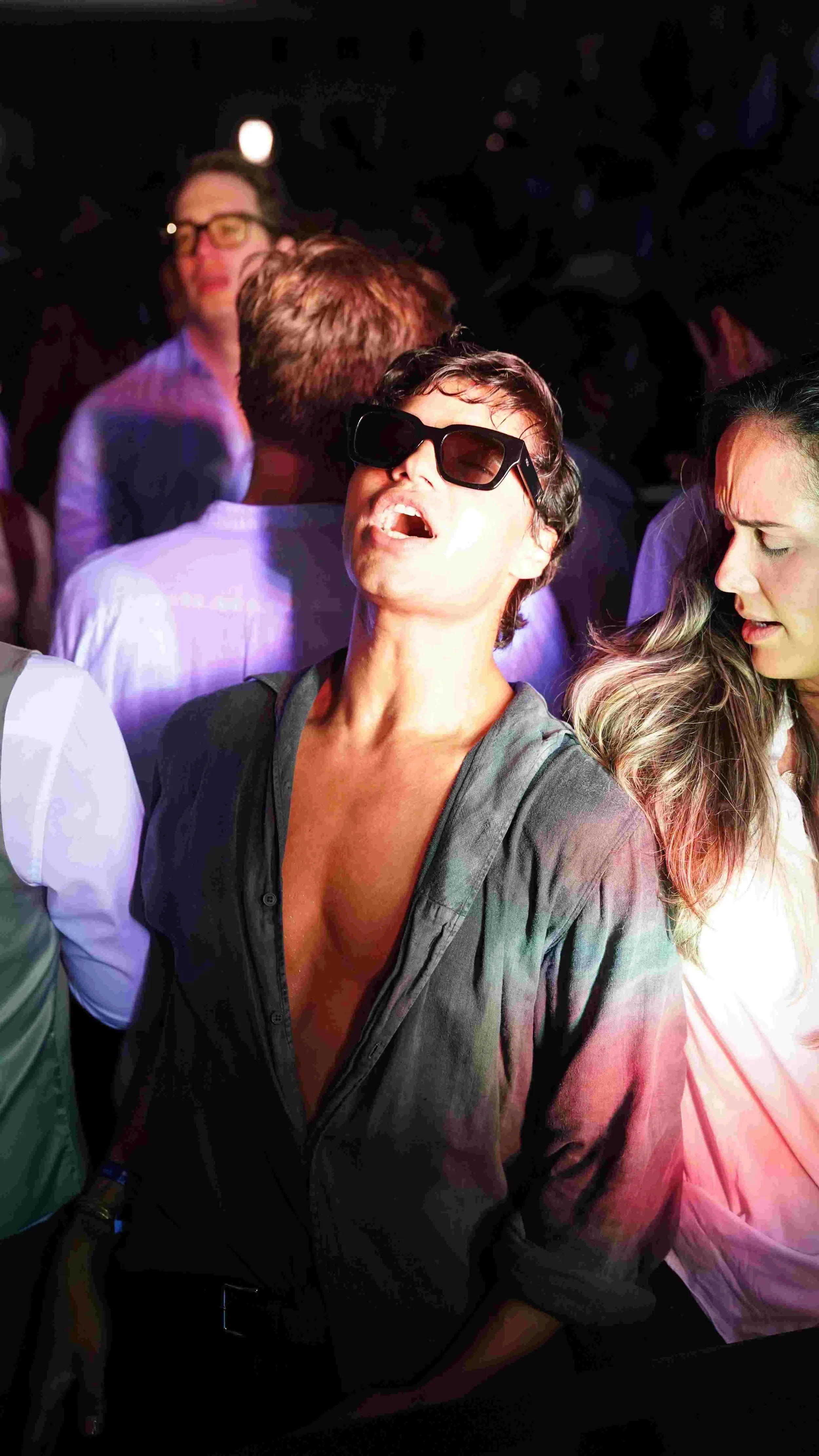 Young woman wearing sunglasses dancing with her mouth open in a crowded party or club with colorful lighting.