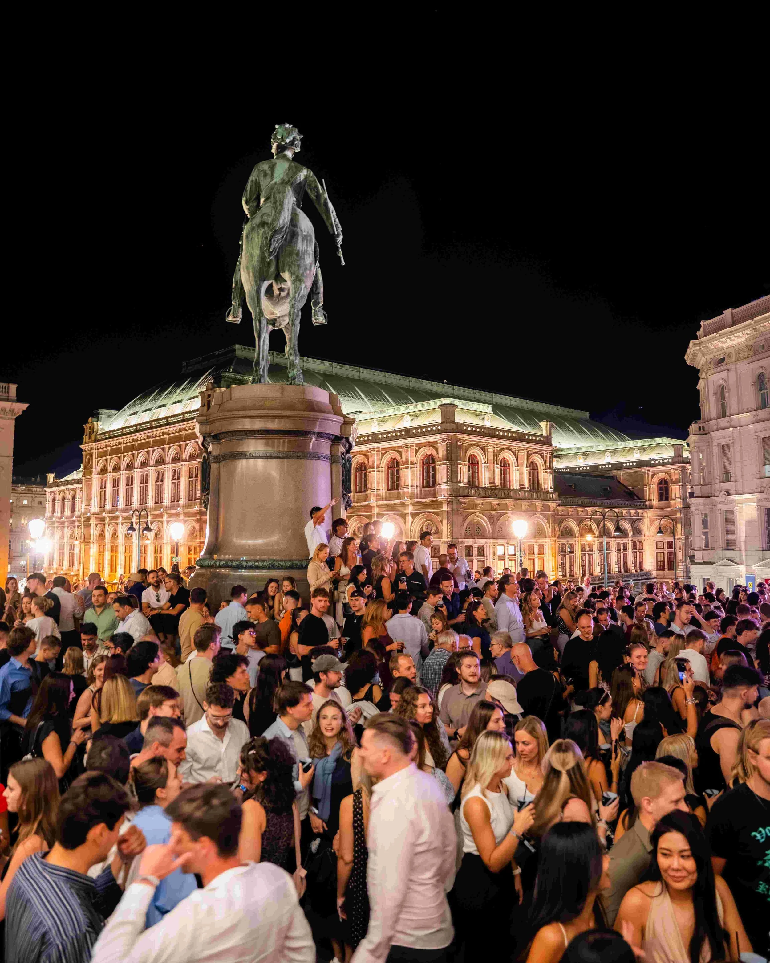 Crowd of people gathered outside a historic European building at night. A large equestrian statue sits atop a prominent pedestal in the foreground. The building is illuminated and has detailed architecture with arched windows and a domed roof.