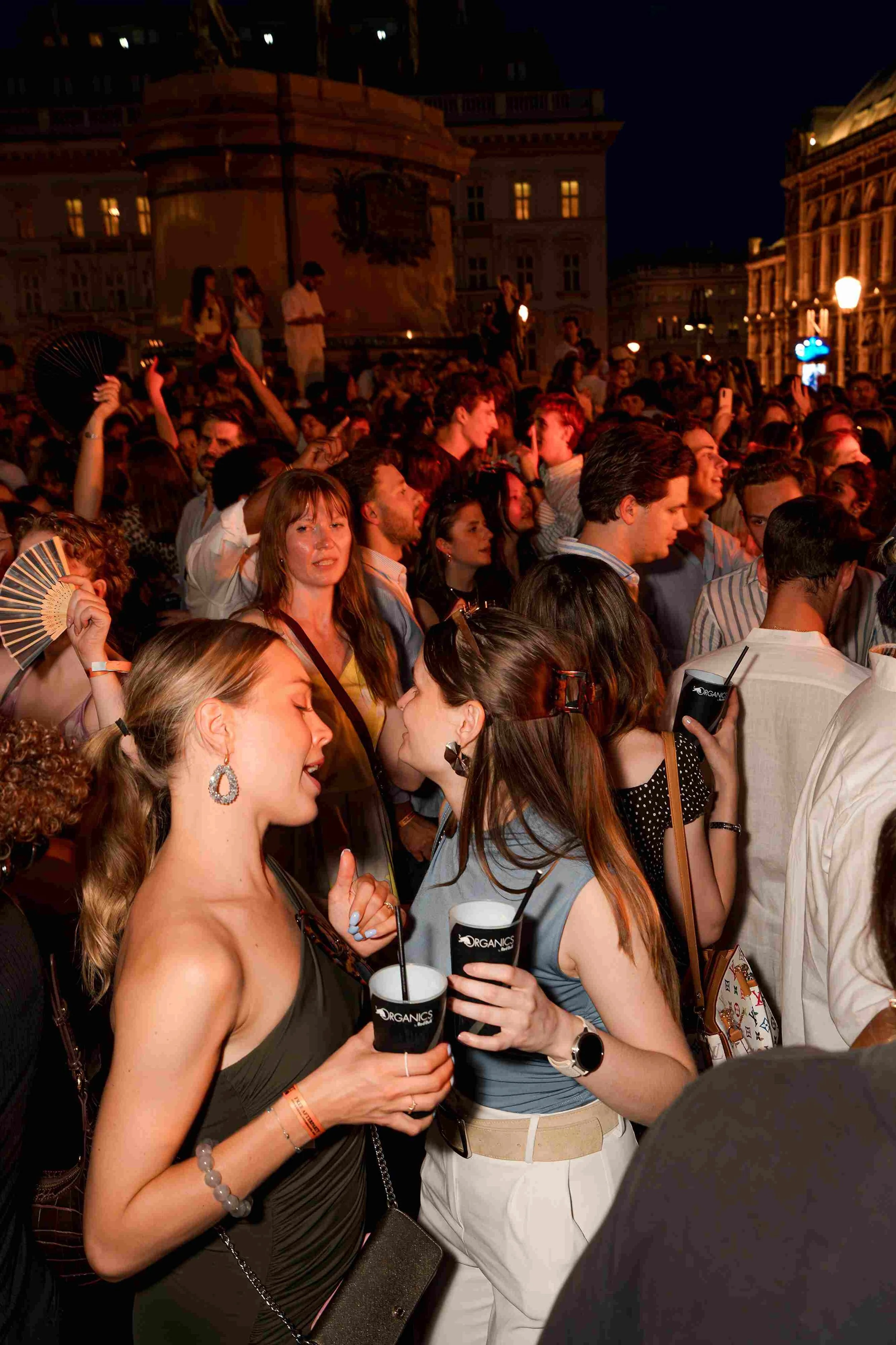 A large crowd of people gathered outdoors at night, some holding drinks and fans, with historic buildings and street lamps in the background.