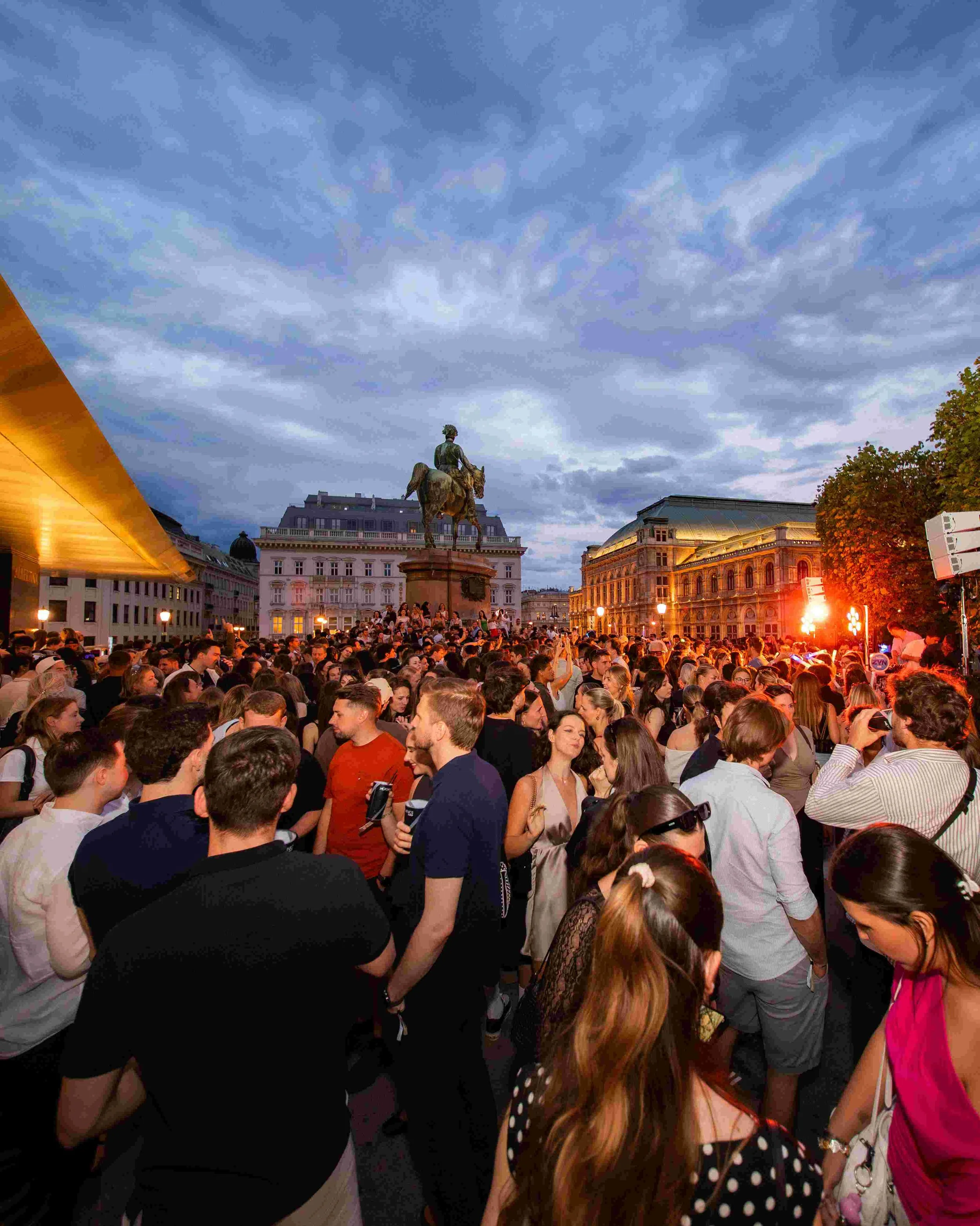 Crowd of people gathered outdoors in an urban setting with historic buildings and a statue of a man on horseback in the background.