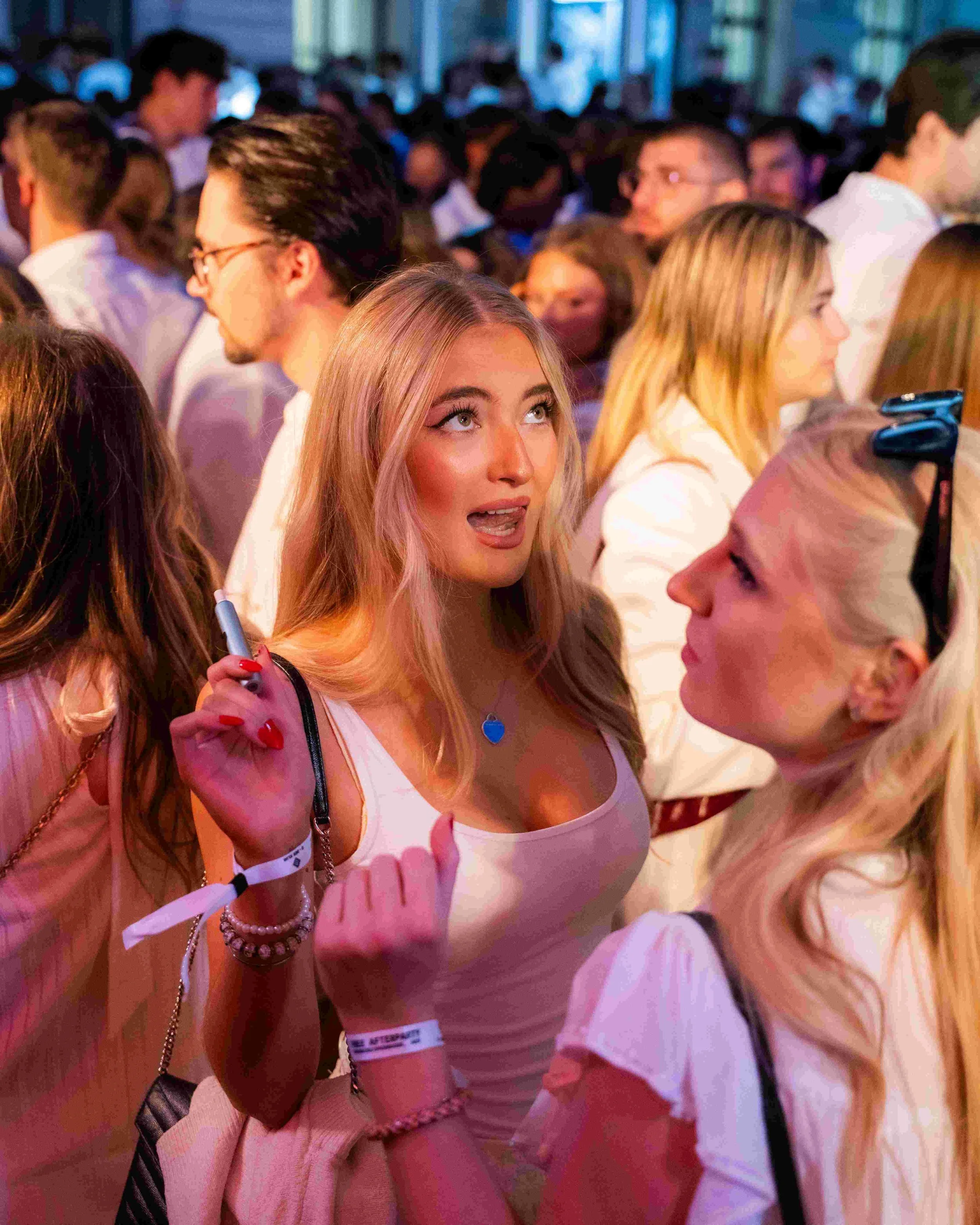 A young woman with blonde hair, wearing a white tank top, is in a crowd at an indoor event, holding a pen and looking upward with an expression of surprise or excitement.