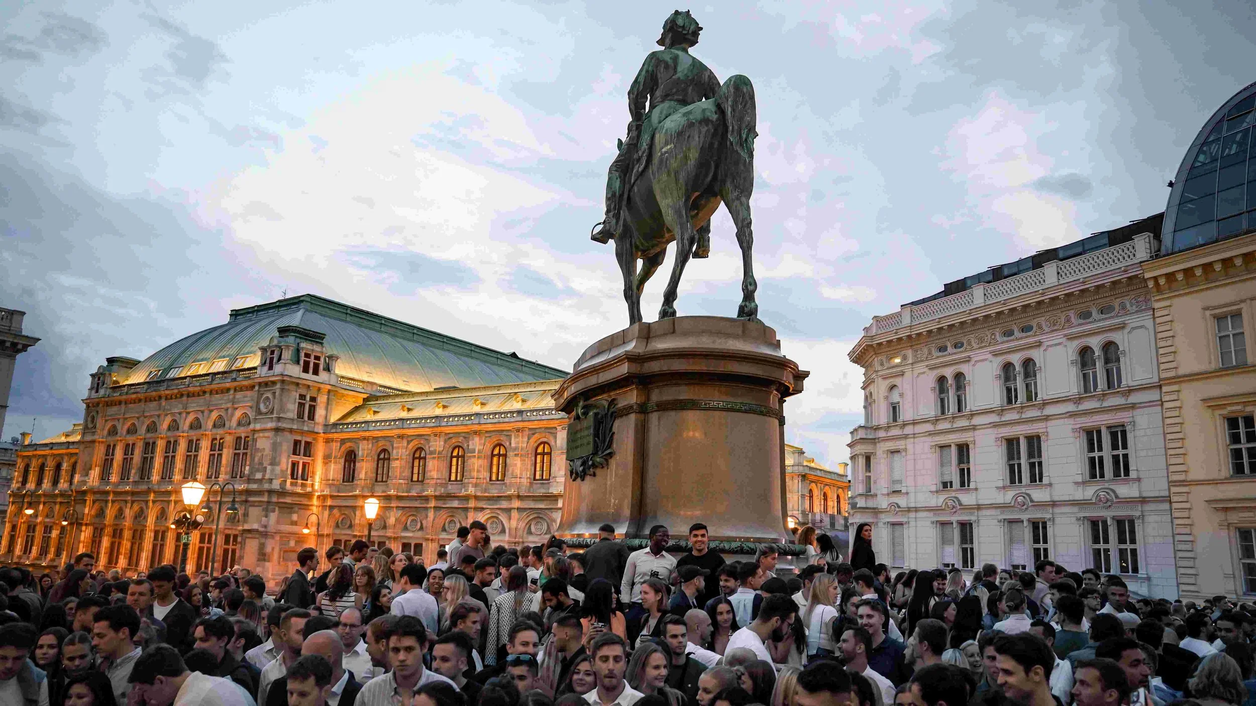 A large crowd gathered around a bronze equestrian statue of a man in a city square during evening, with historic buildings and cloudy sky in the background.