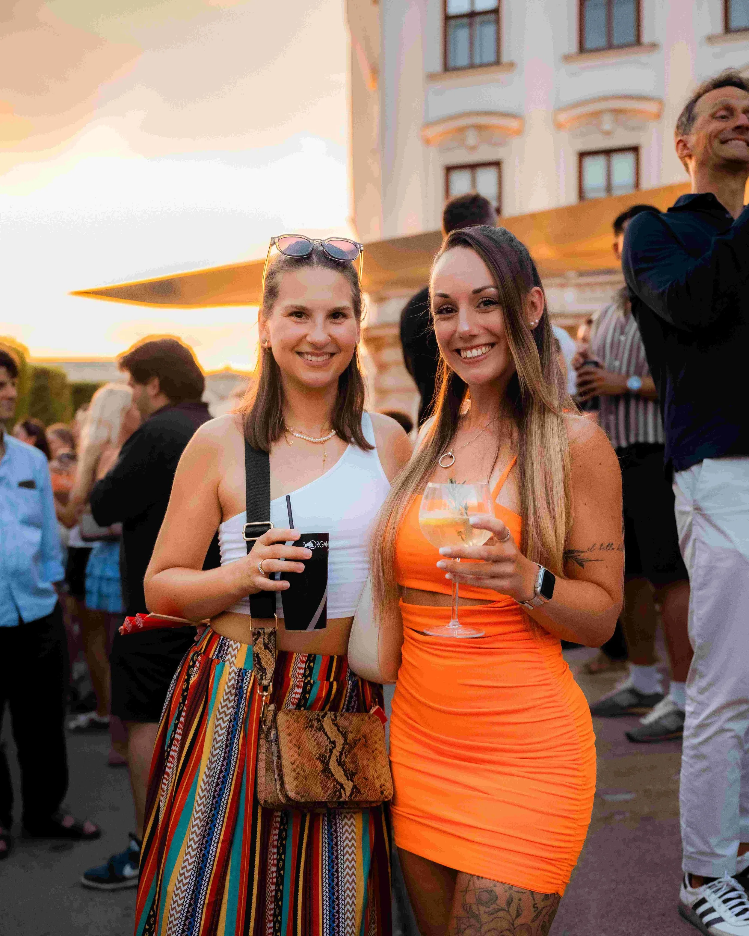 Two smiling women at a social outdoor event during sunset, one holding a drink and the other with sunglasses on her head, with a crowd of people, a building, and a colorful sky in the background.