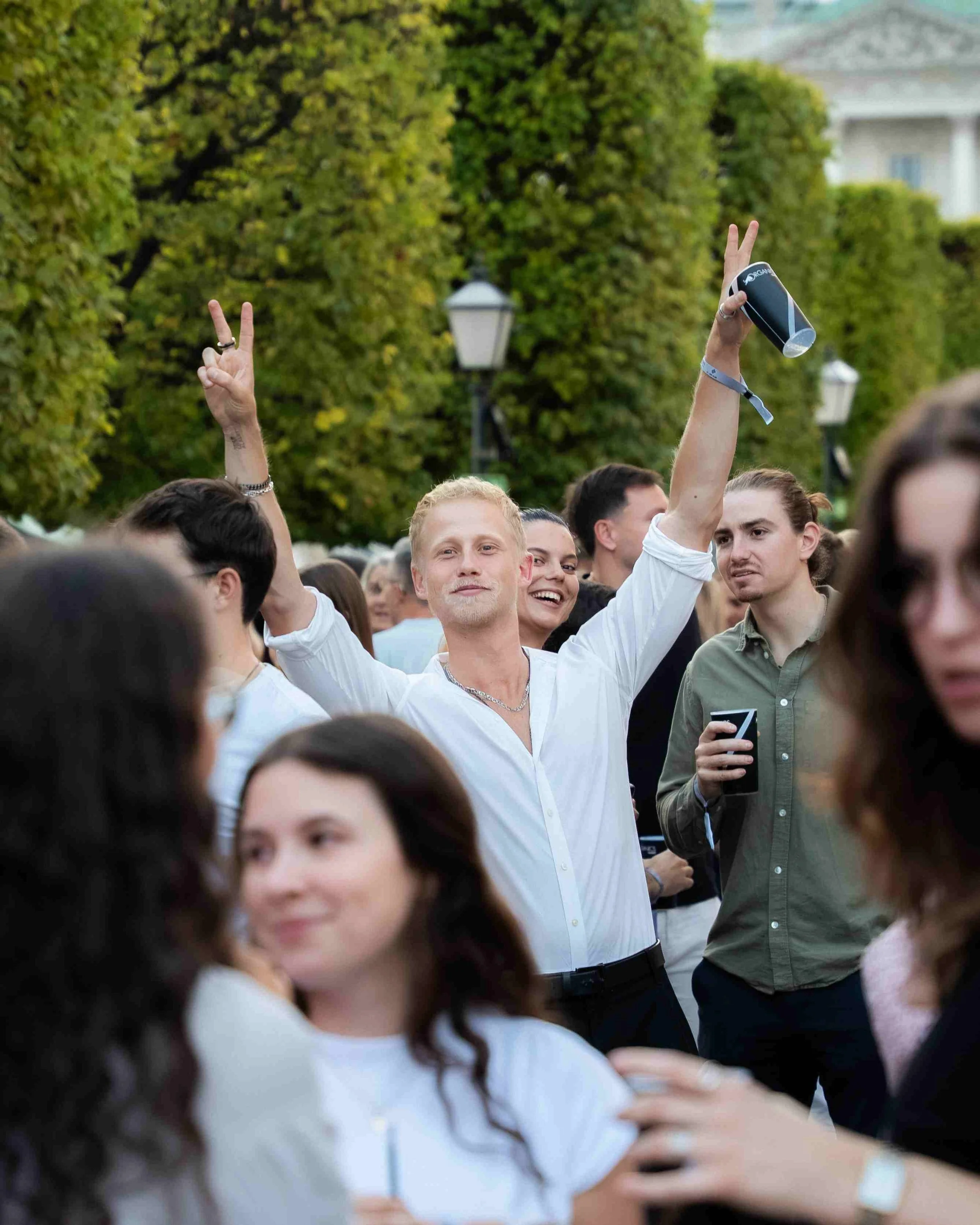 A group of people gathered outdoors, some making peace signs with their hands. There are trees and a building in the background.