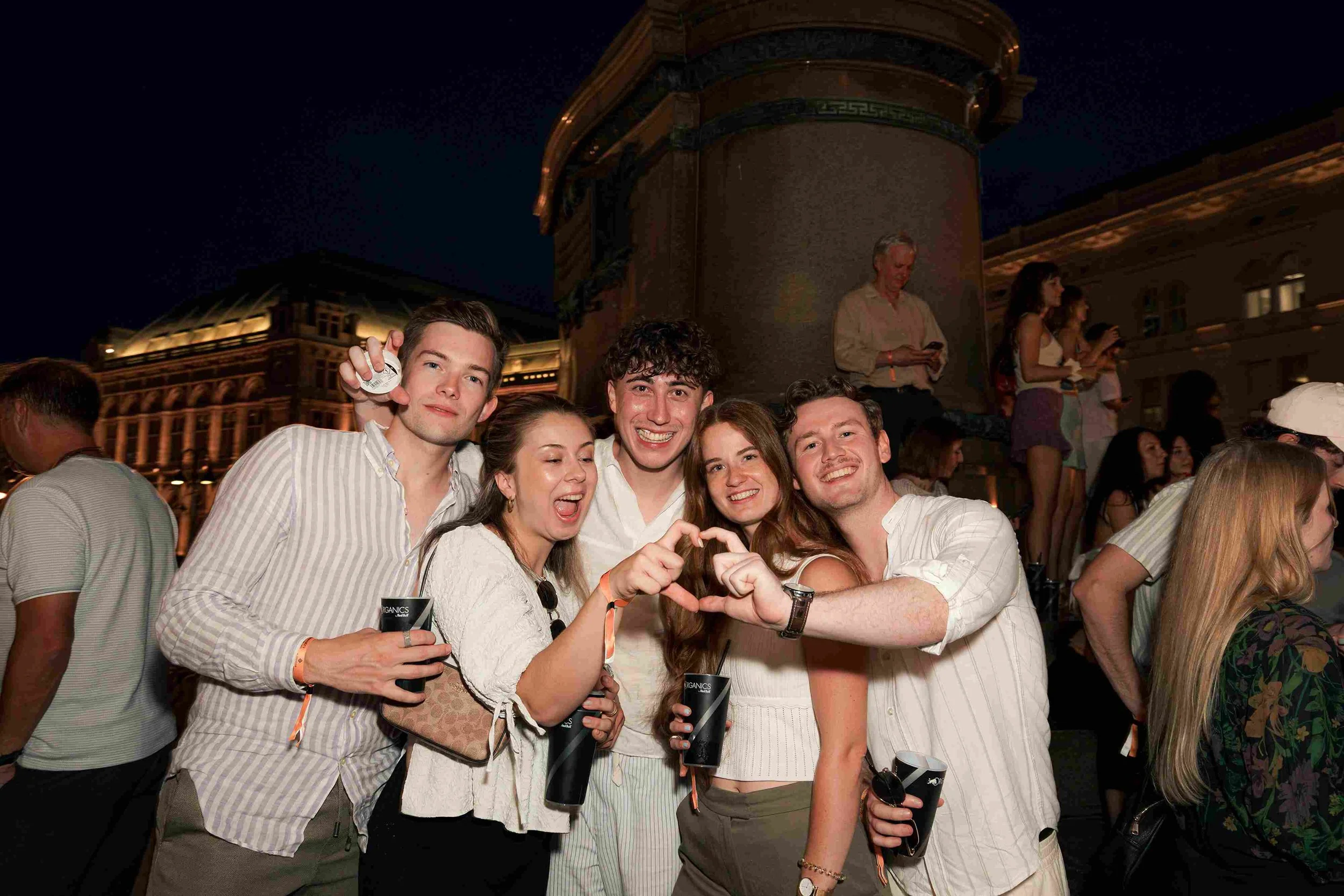 Group of young adults at a nighttime outdoor event, smiling, some holding drinks, in front of historic buildings and a dark sky.