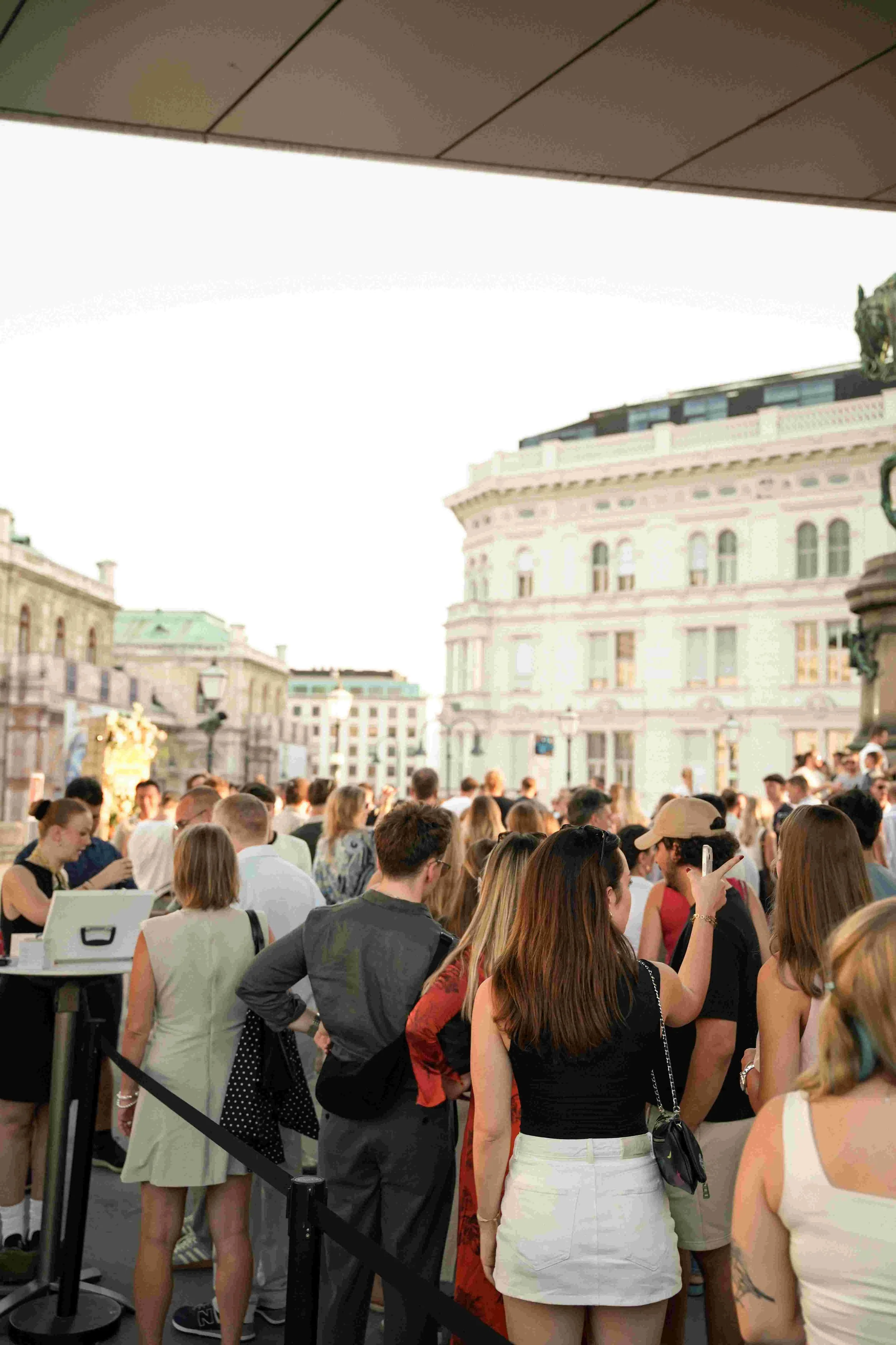 A large crowd of people standing in line outdoors in front of historic buildings, with some using their phones, under a partial roof overhang.