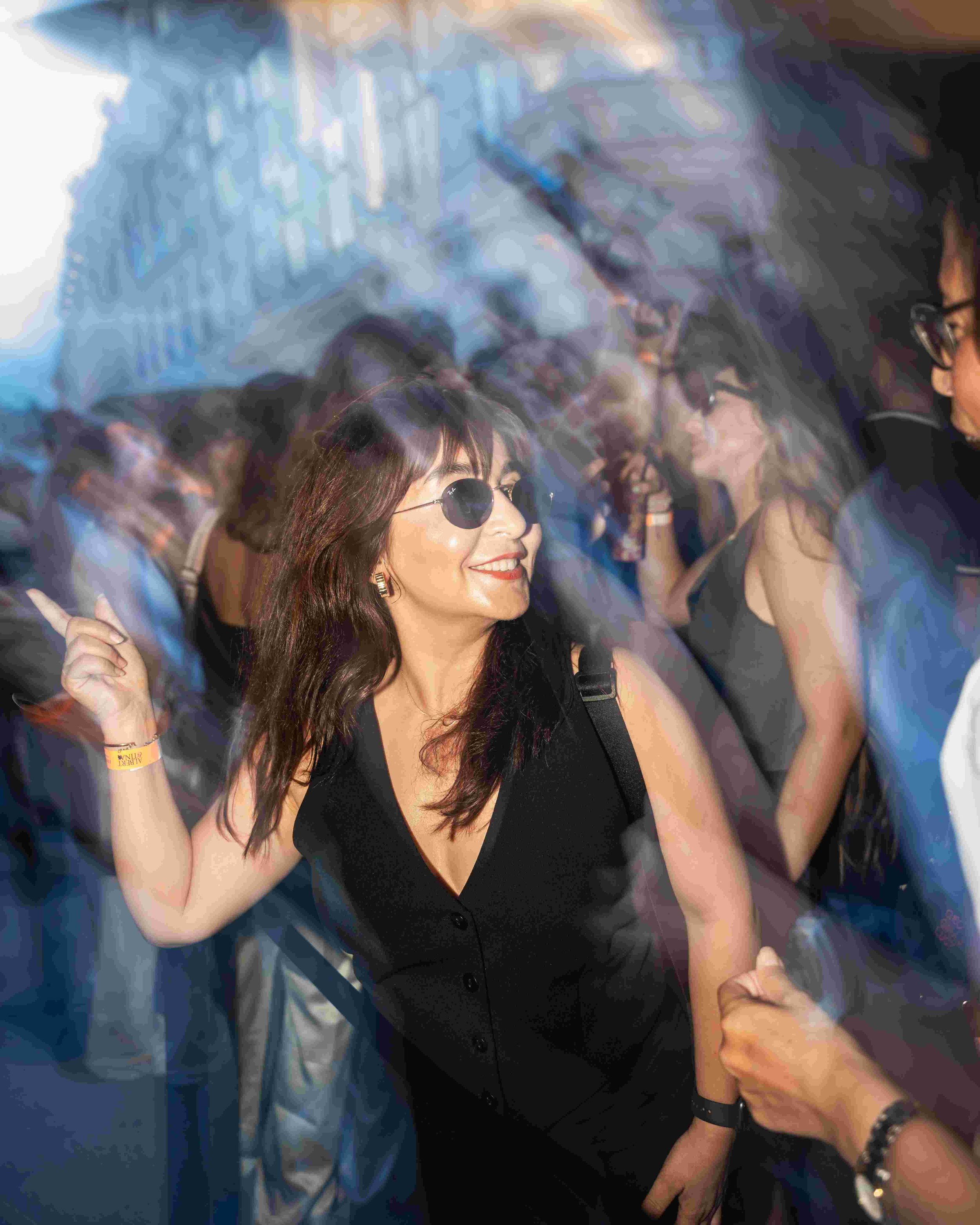 A woman with long brown hair, wearing sunglasses and a black dress, smiling and holding hands with a person whose arm is visible. The woman appears to be at a crowded event or celebration, and the atmosphere seems lively with colorful lights.
