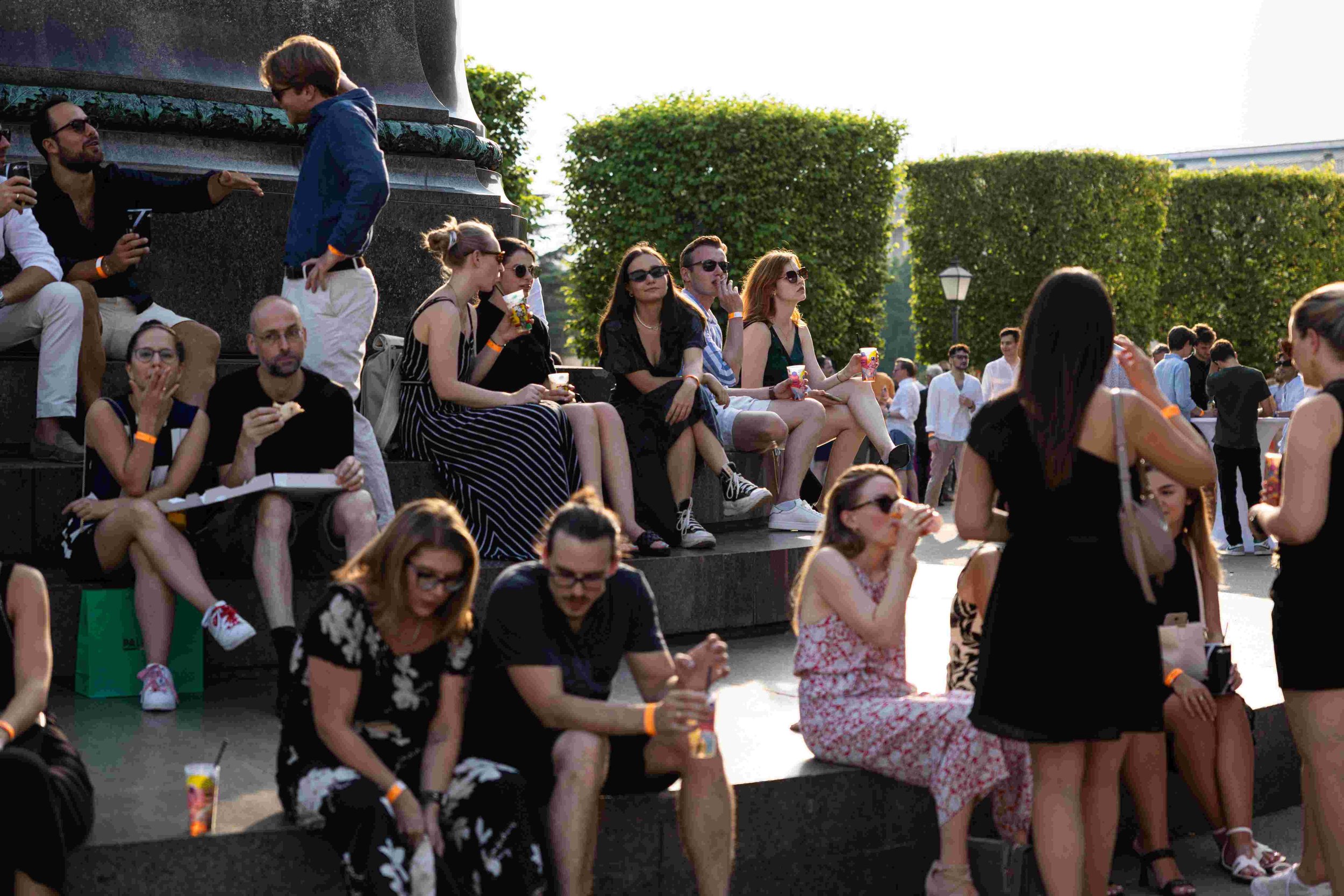 A group of people sitting on outdoor steps at a social event, some holding drinks and food, with sunlight shining on them and greenery in the background.