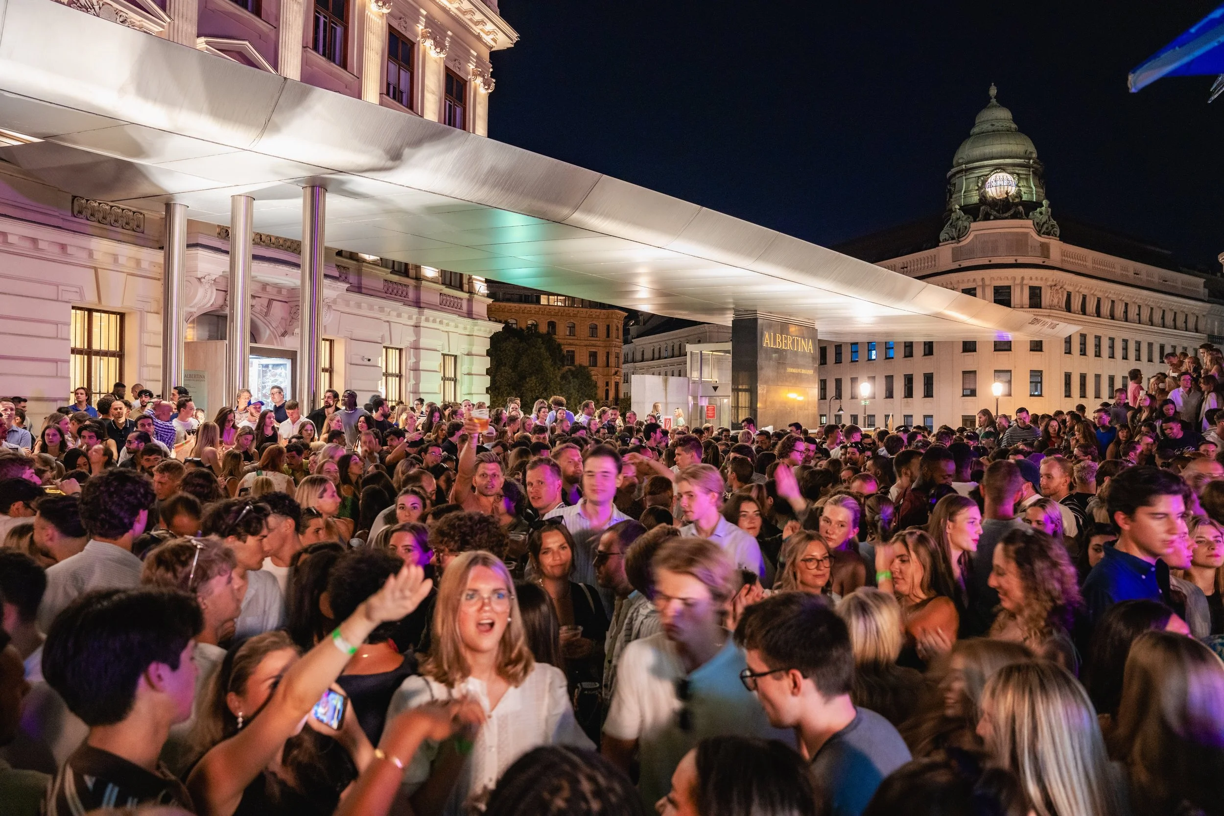 Crowd of people gathered outside a building at night, with some holding drinks and taking photos, under lit architecture and a dark sky.