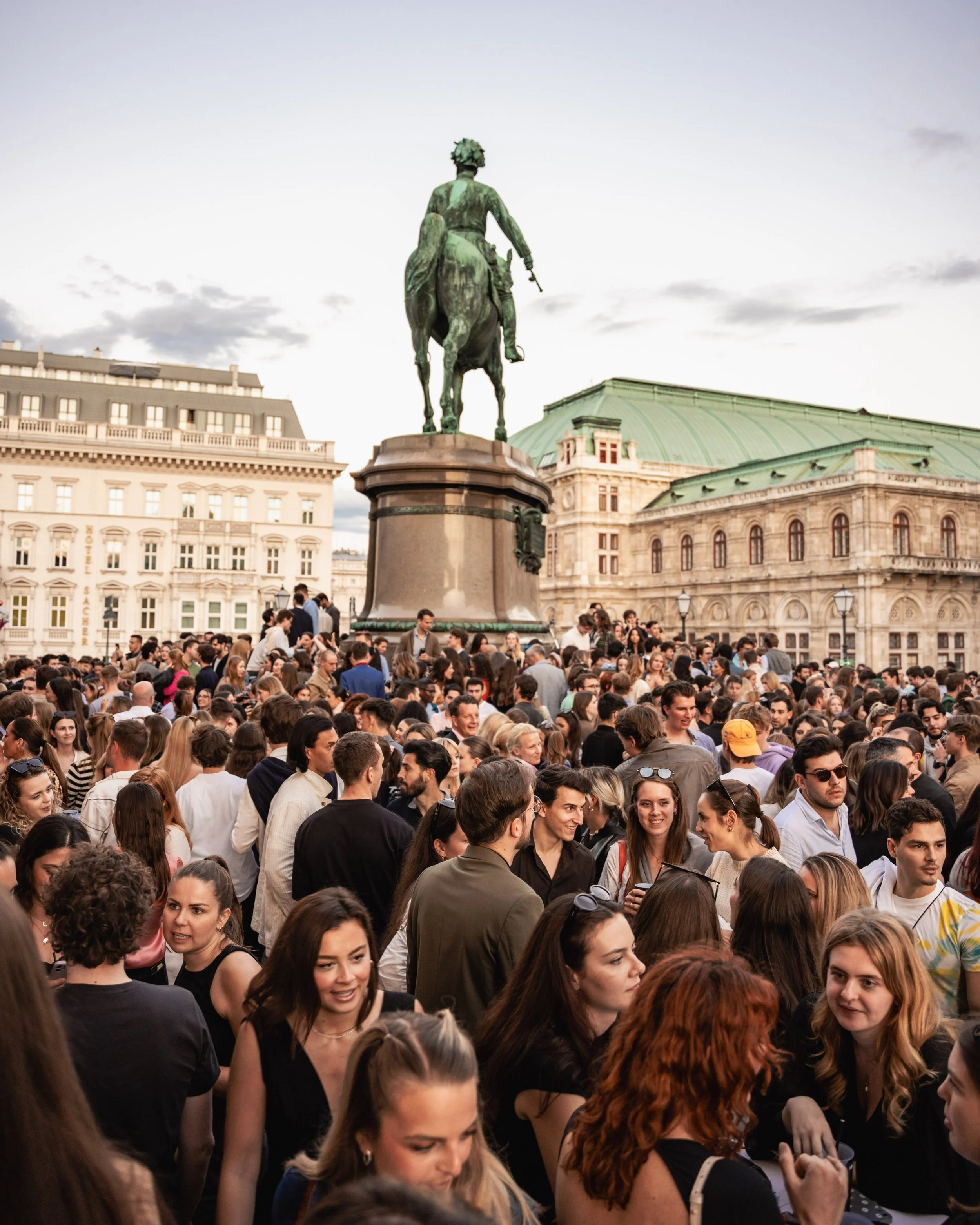 Crowd of people gathered outdoors in front of a historic building with a green roof, situated behind an equestrian statue of a man on a horse.