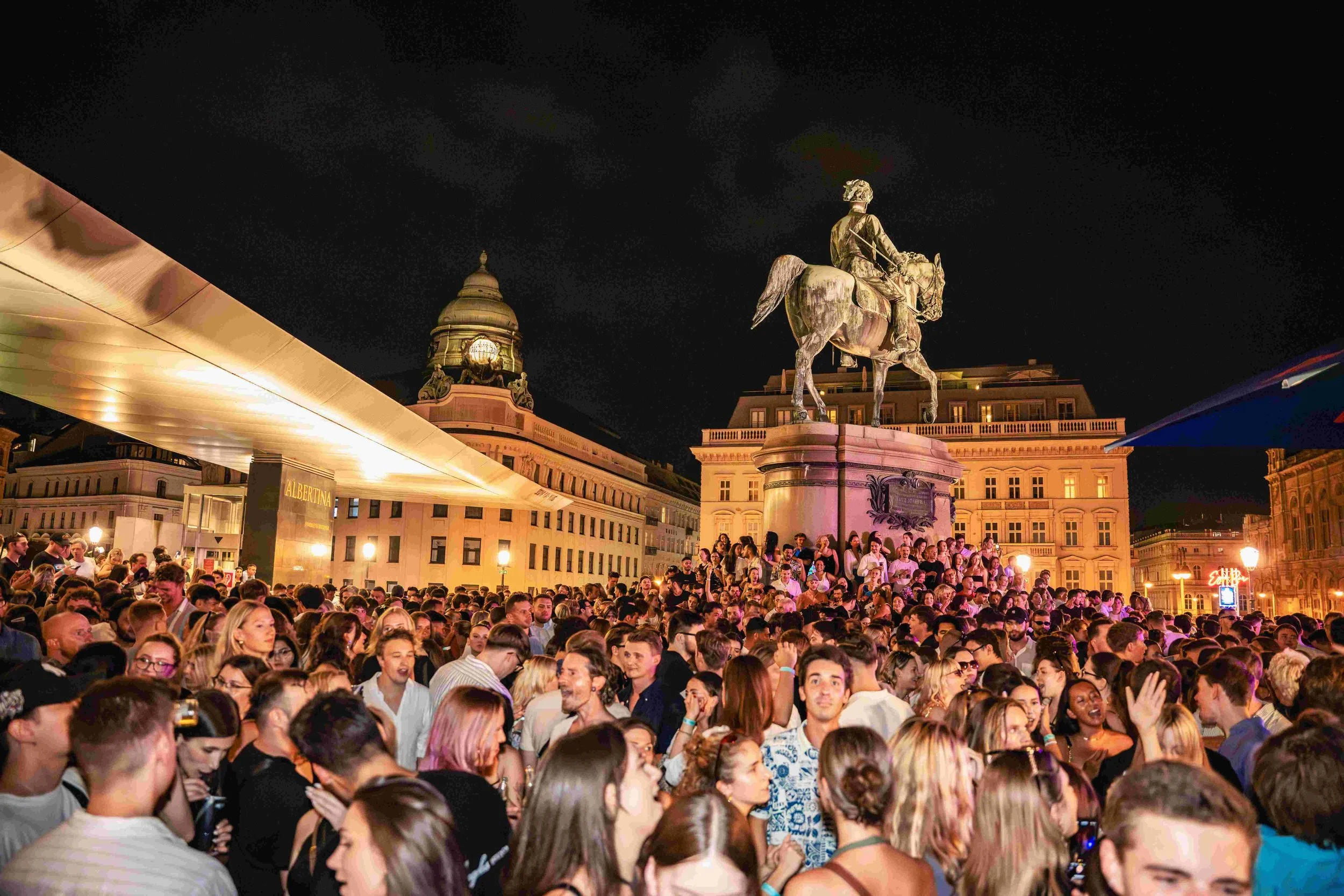 A large crowd of people gathered outdoors at night, with historical buildings and an equestrian statue in the background, illuminated by artificial lights.