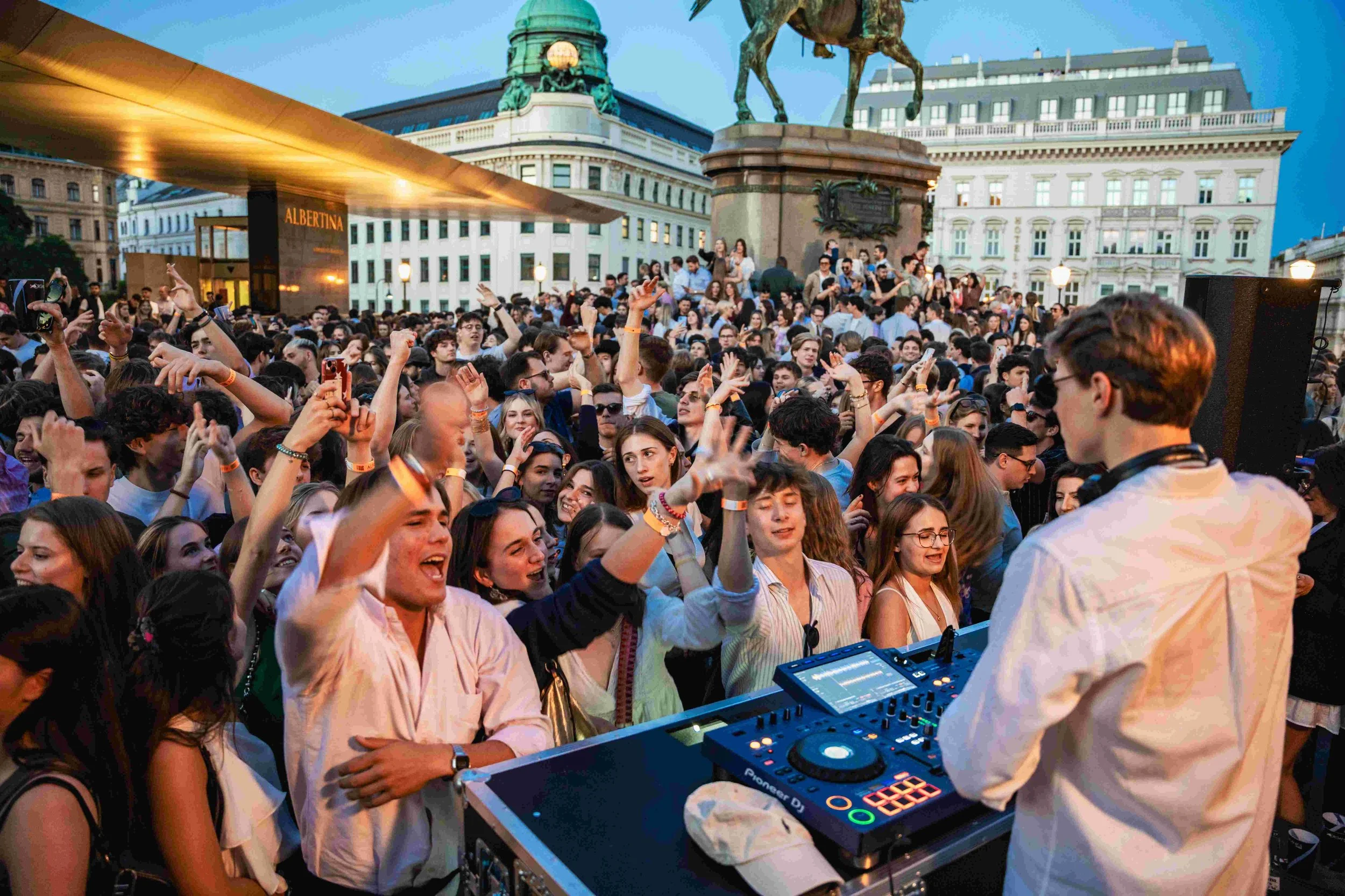 Crowd of young people at outdoor music event with DJ in foreground, city buildings, and monument in background.