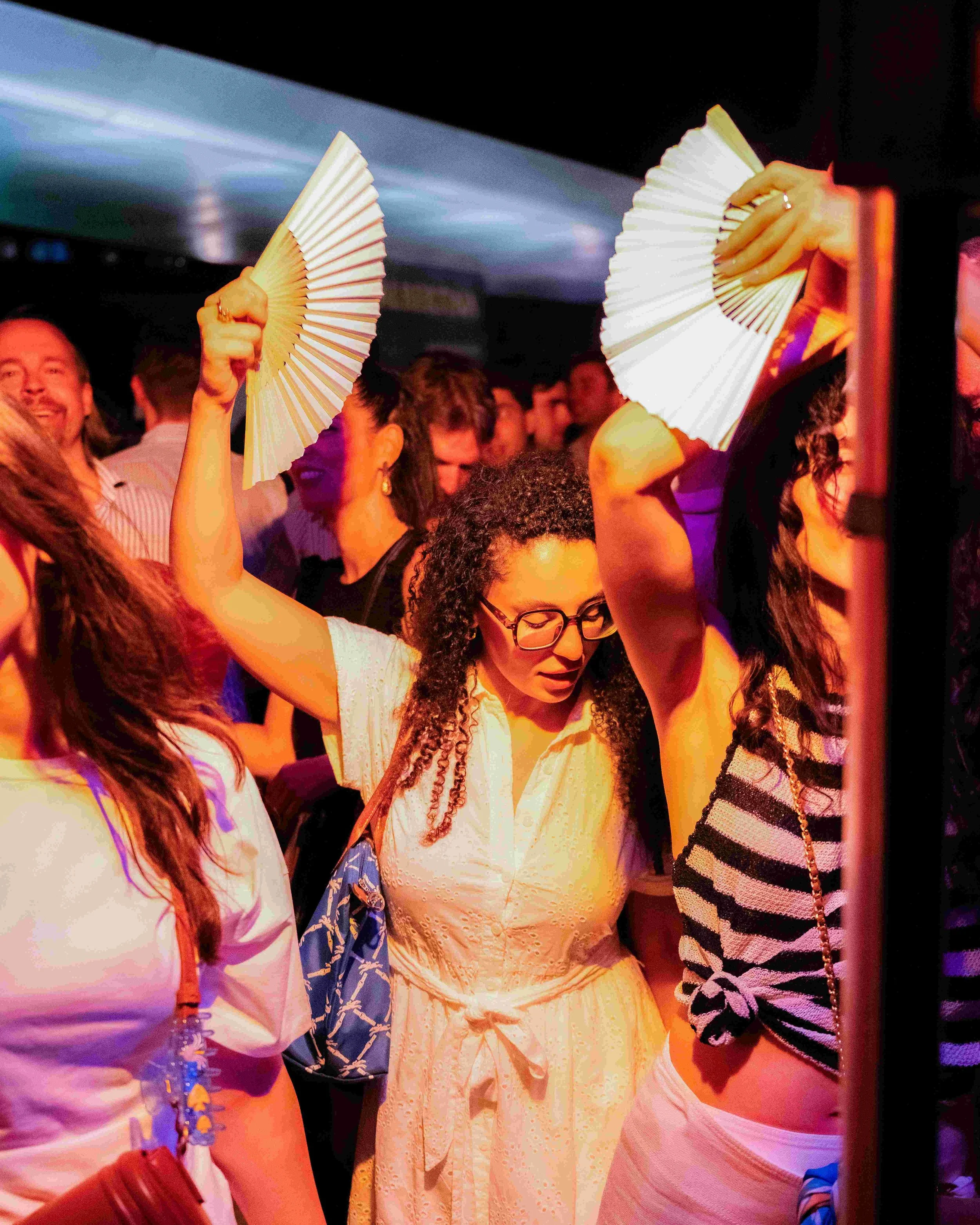 People at a party or festive event, with some holding folding fans. The scene has warm lighting and a crowd of individuals engaged in conversation or enjoying the event.