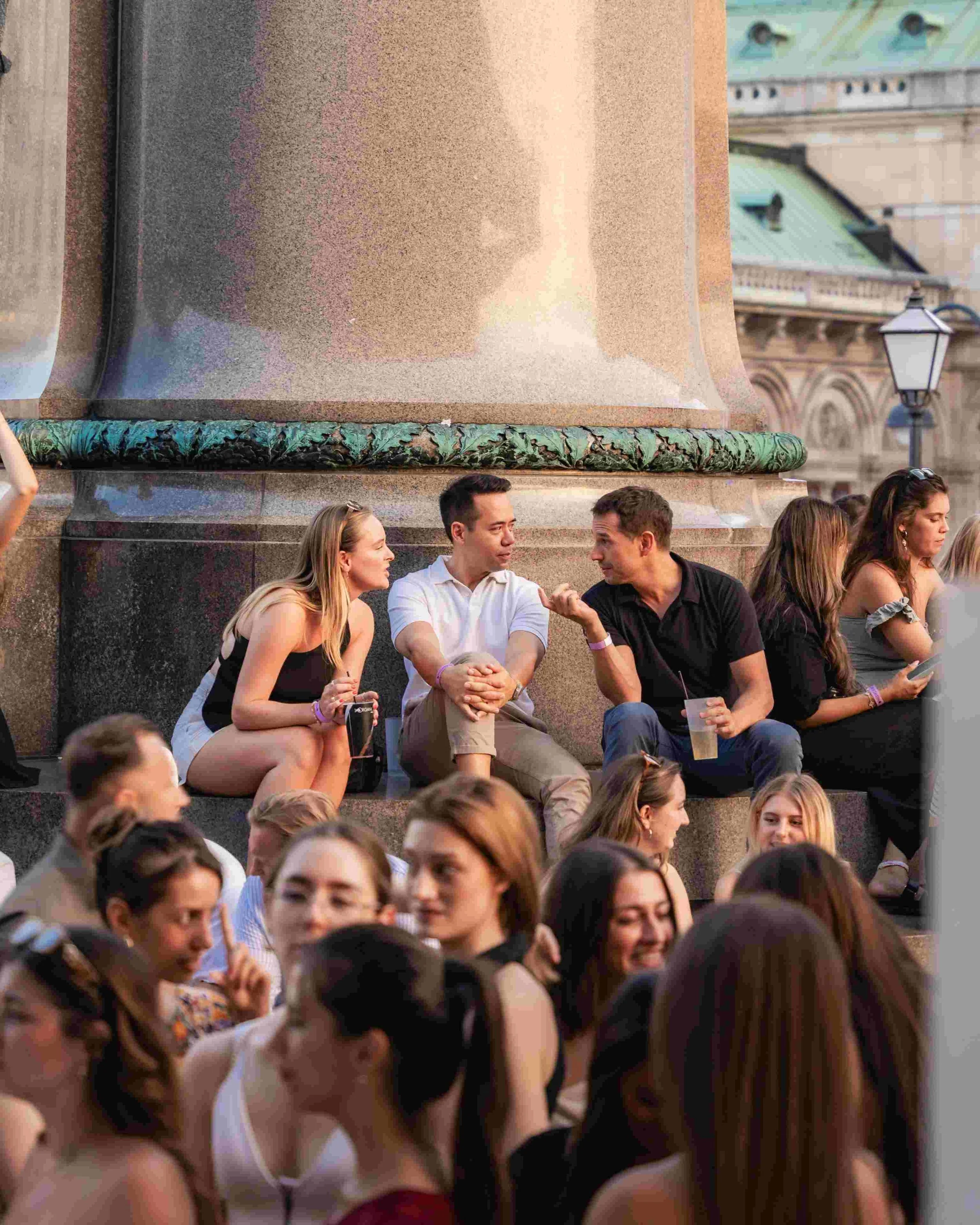 A group of people sitting on steps outdoors, engaged in conversation, with a large stone structure and a streetlamp in the background.