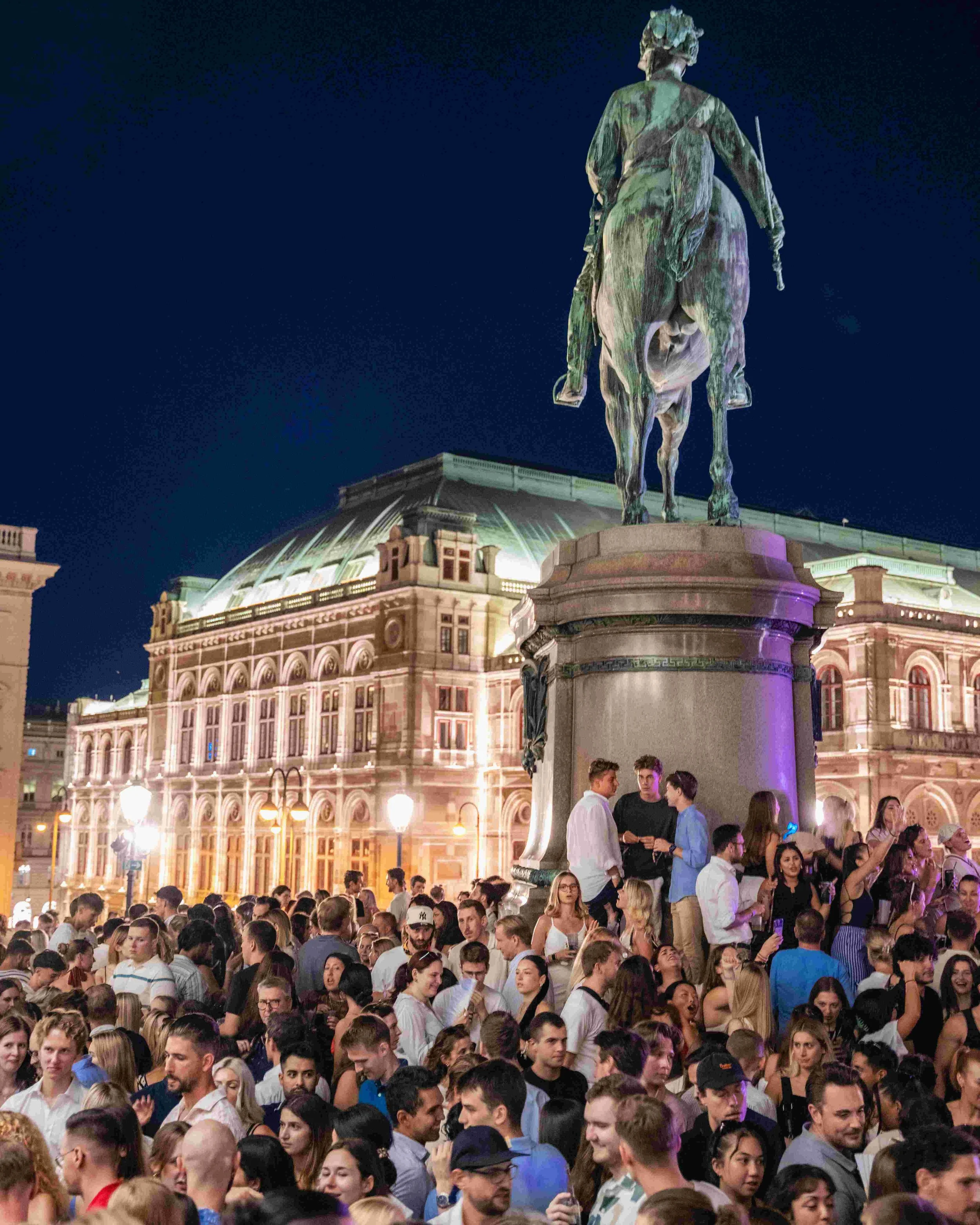Nighttime scene of a large crowd gathered in front of an ornate, historic building with a glowing green dome. A prominent statue of a man on horseback stands in the foreground, partially illuminated.