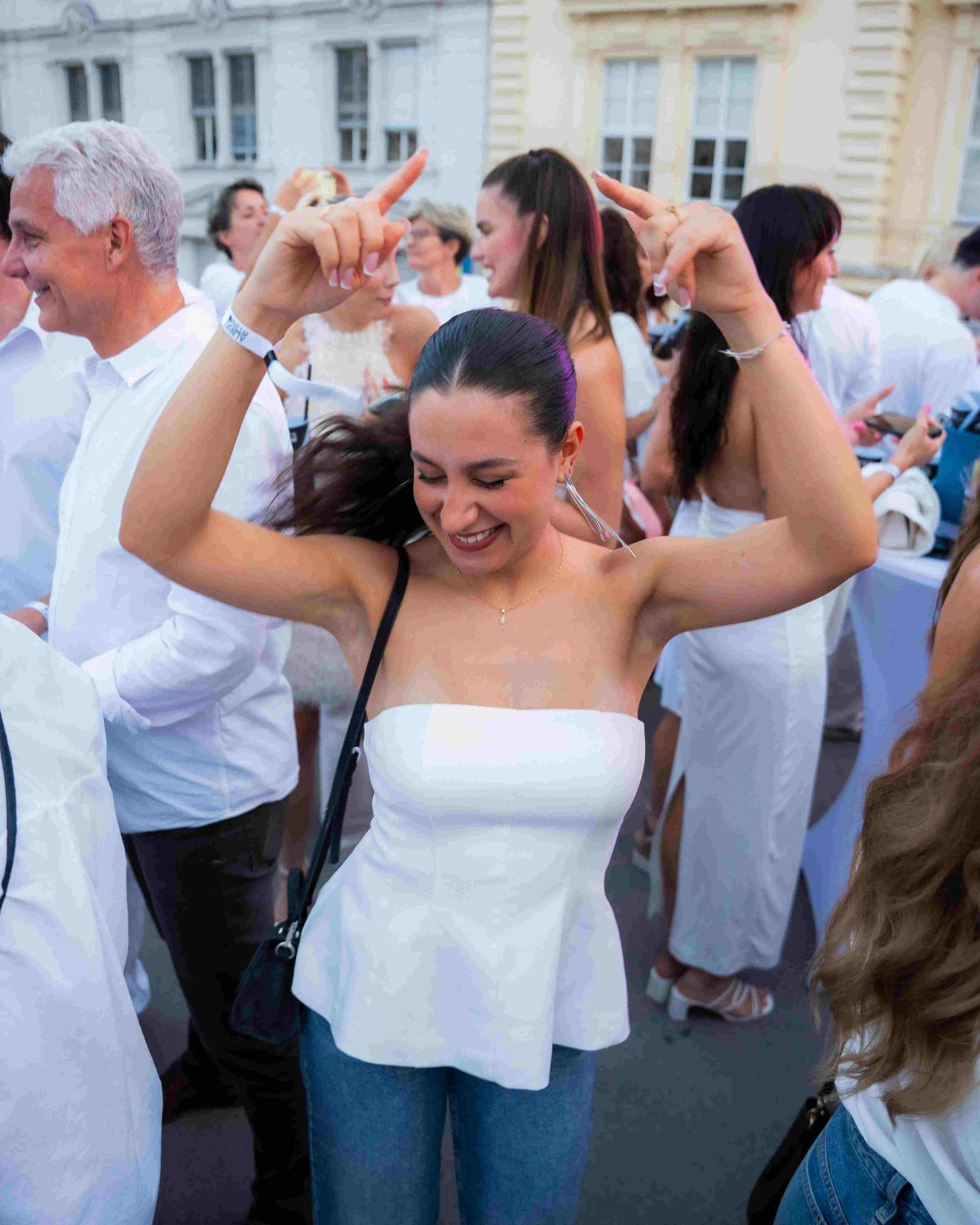 A group of people at an outdoor event, dressed in white, are dancing and enjoying themselves. The focus is on a young woman with dark hair, smiling and dancing with her arms raised.