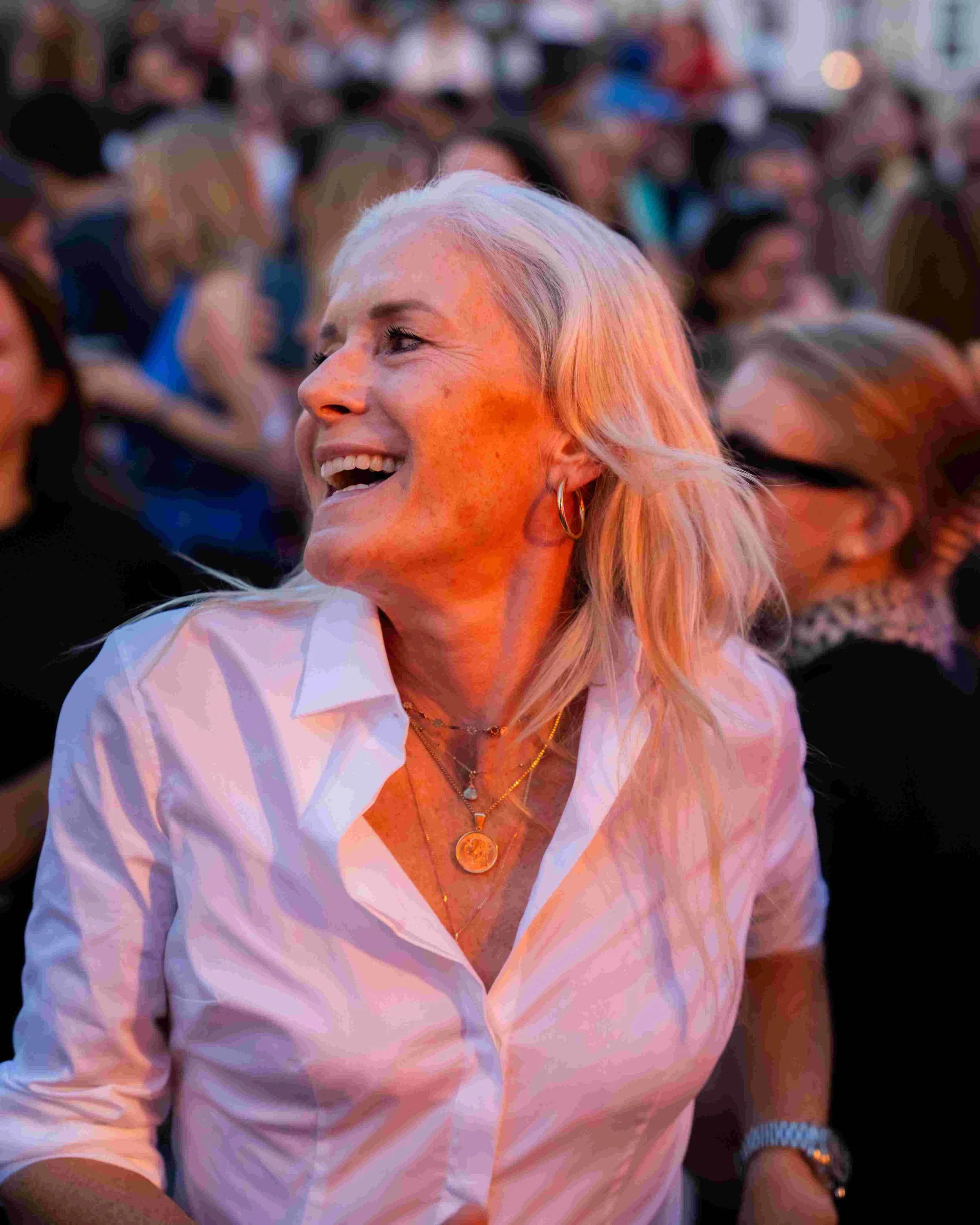 A woman with long blonde hair smiling and looking to her right, wearing a white shirt, layered necklaces, and gold hoop earrings, in a crowd at what appears to be a lively event or gathering.