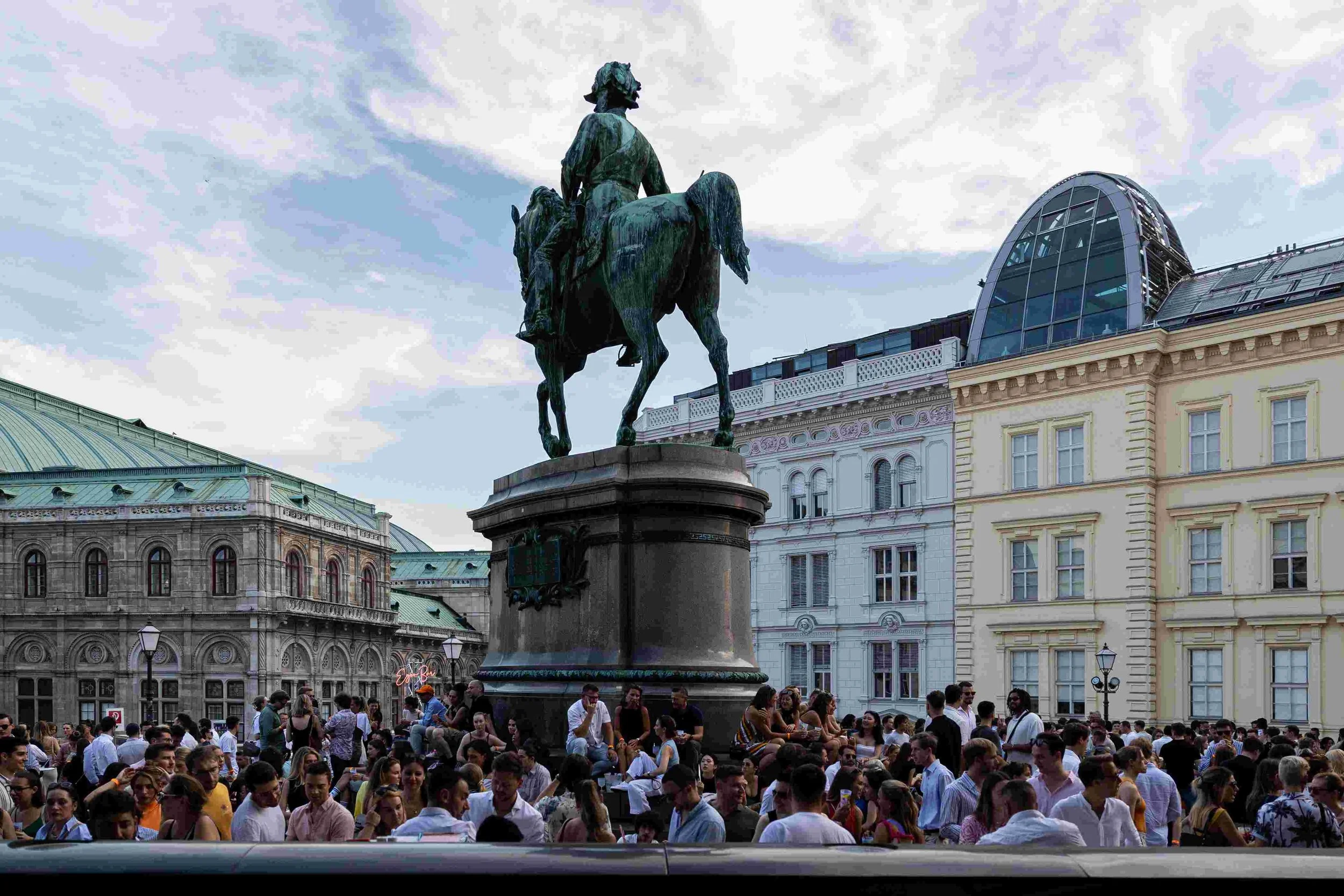 A large crowd of people gathered in a city square with an equestrian statue of a man on horseback at the center. The background shows historic and modern buildings, with a cloudy sky overhead.