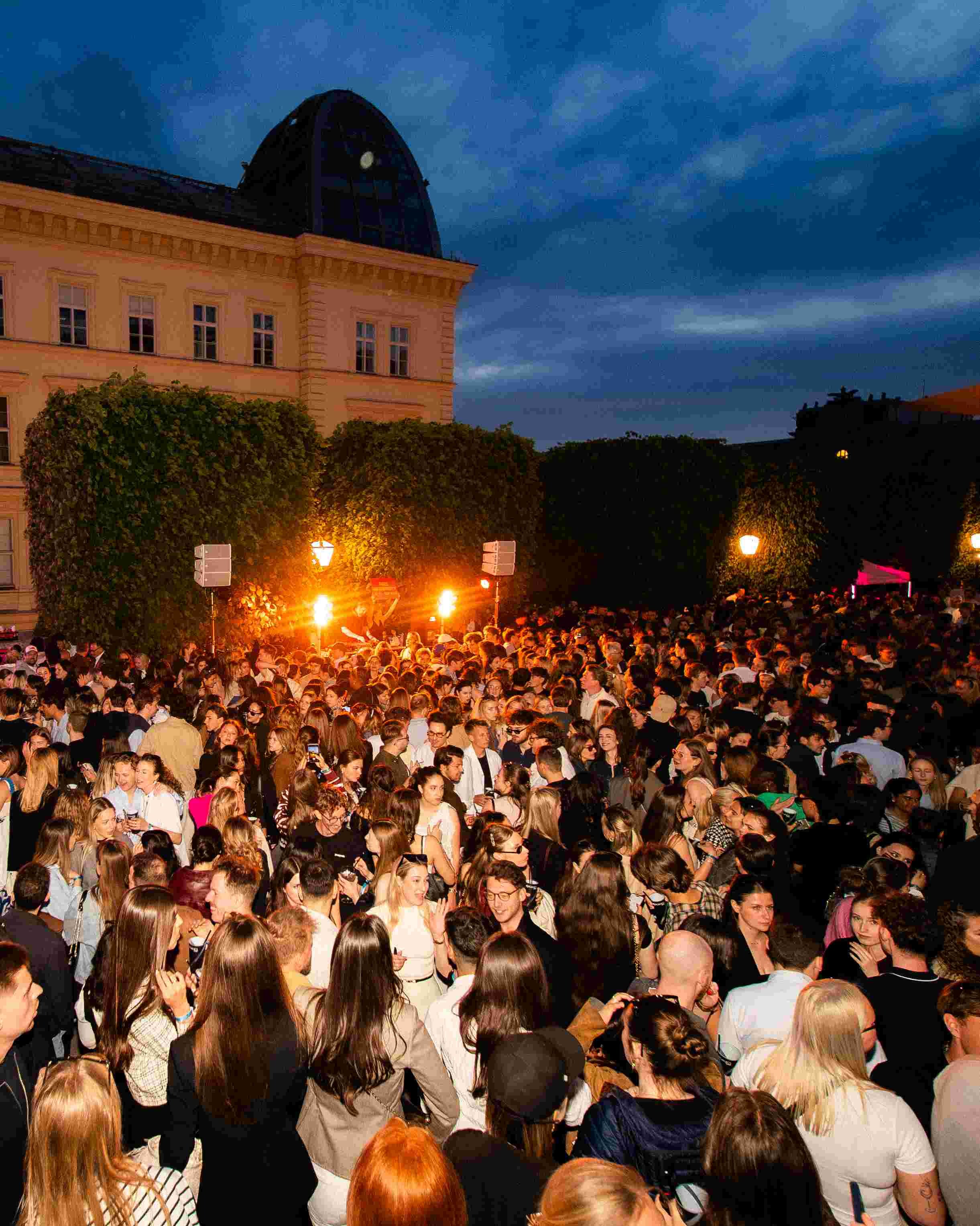 A large crowd of people gathered outdoors during evening, with many facing forward or chatting, under a dark, cloudy sky, with street lamps illuminating the scene and a historic building with a glass dome in the background.