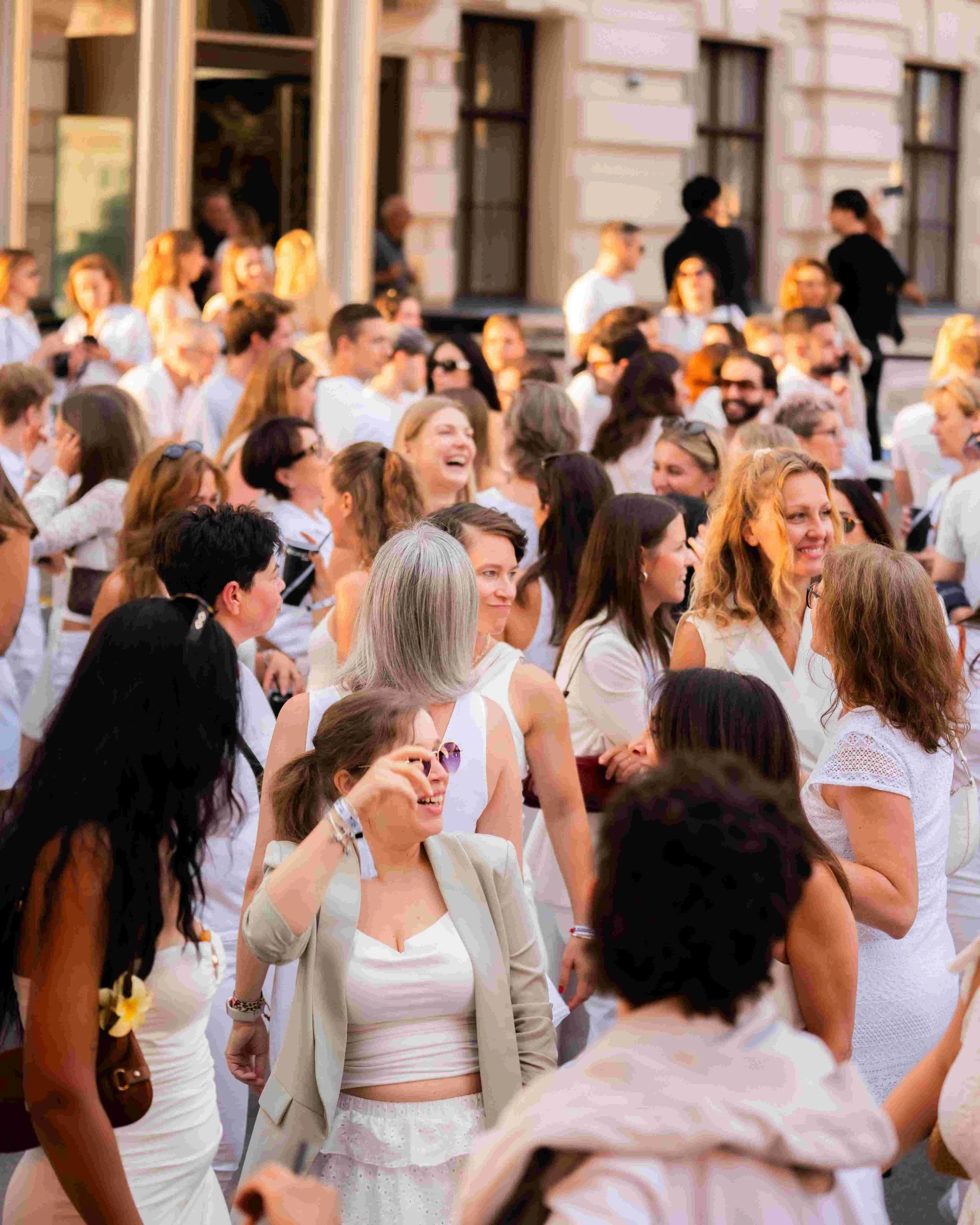 Crowd of people dressed in white, smiling and talking outside a building with large windows.