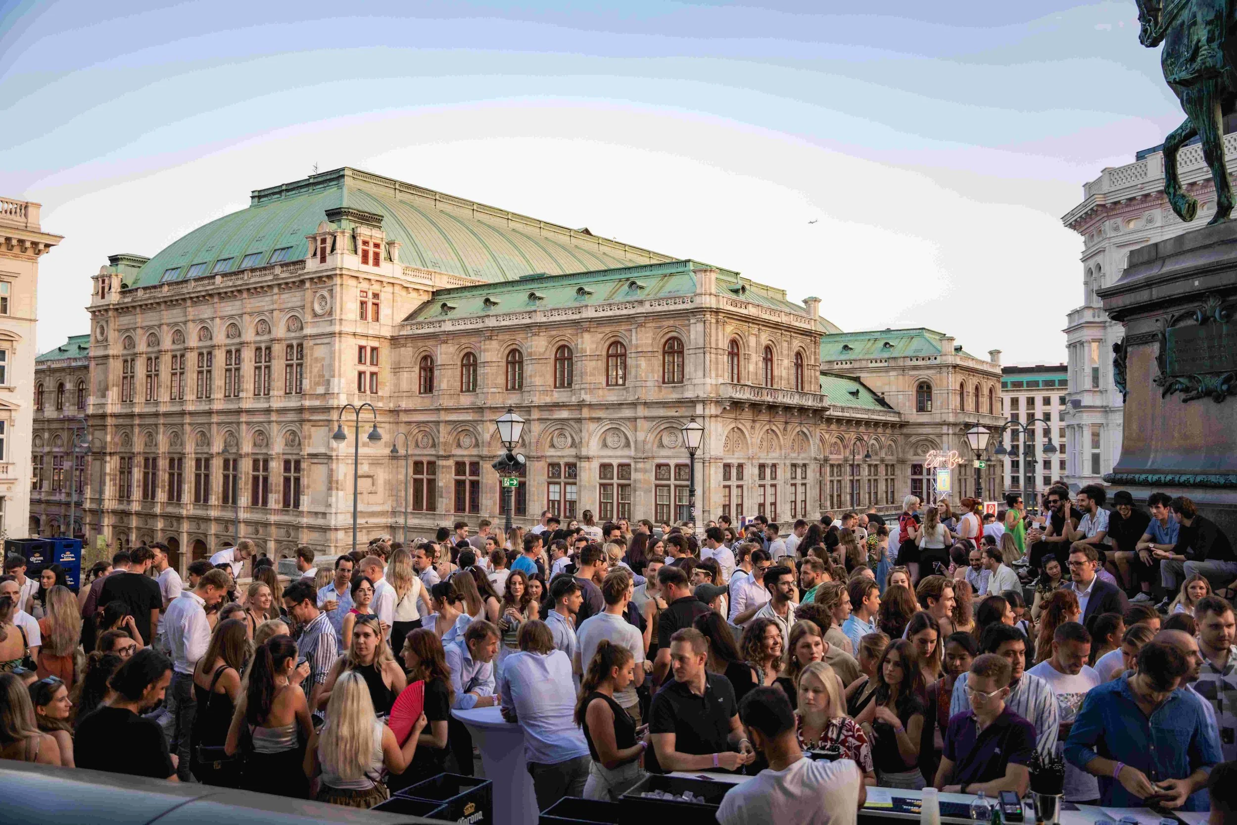 A large crowd of people gathered outdoors in front of historic buildings with green rooftops during the daytime.