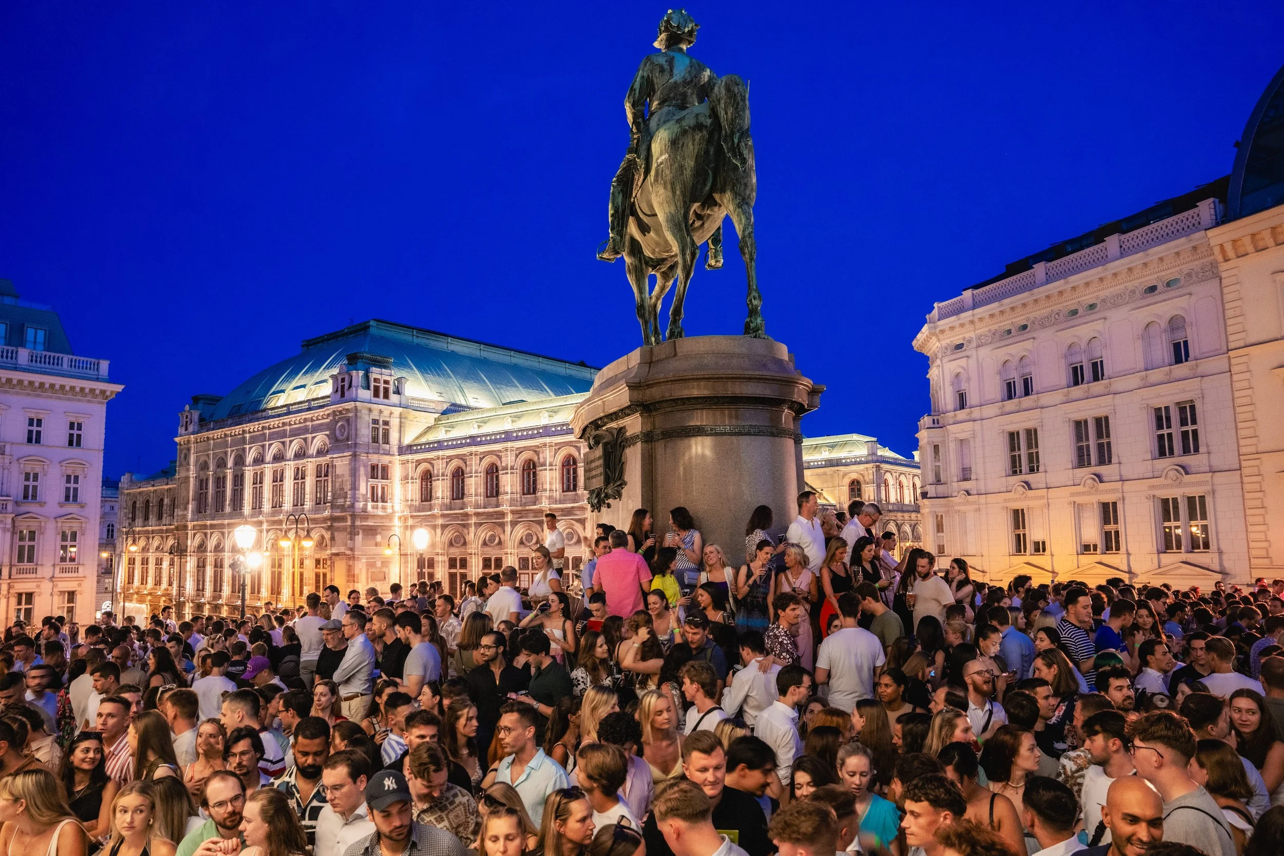 Crowd of people gathered outdoors at night with historic buildings and an equestrian statue in the background, illuminated by streetlights and city lighting.