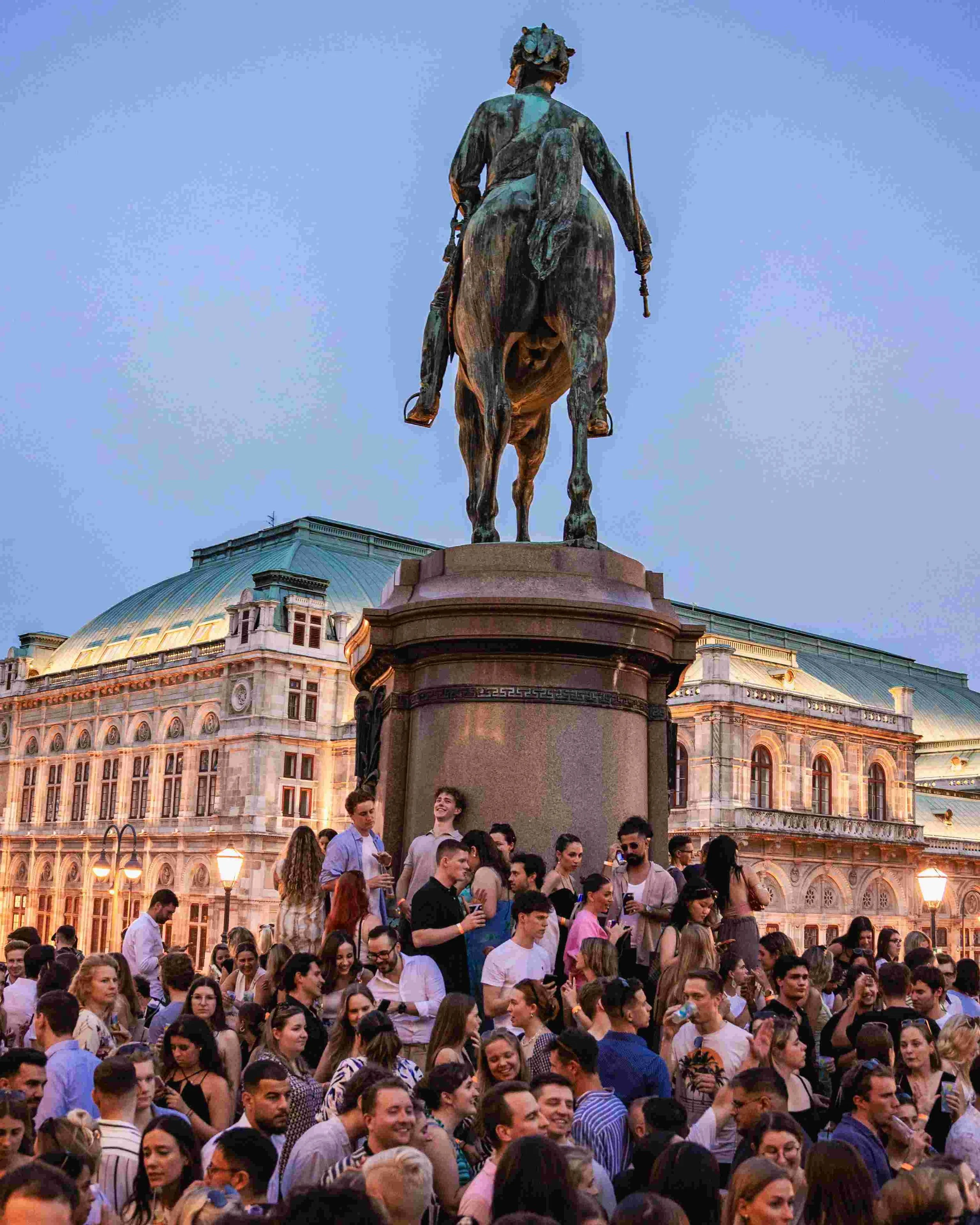 A crowd of people gathered in front of a large equestrian statue during early evening. The statue depicts a man riding a horse, positioned on a pedestal, with an historic building featuring arched windows and a dome roof in the background.