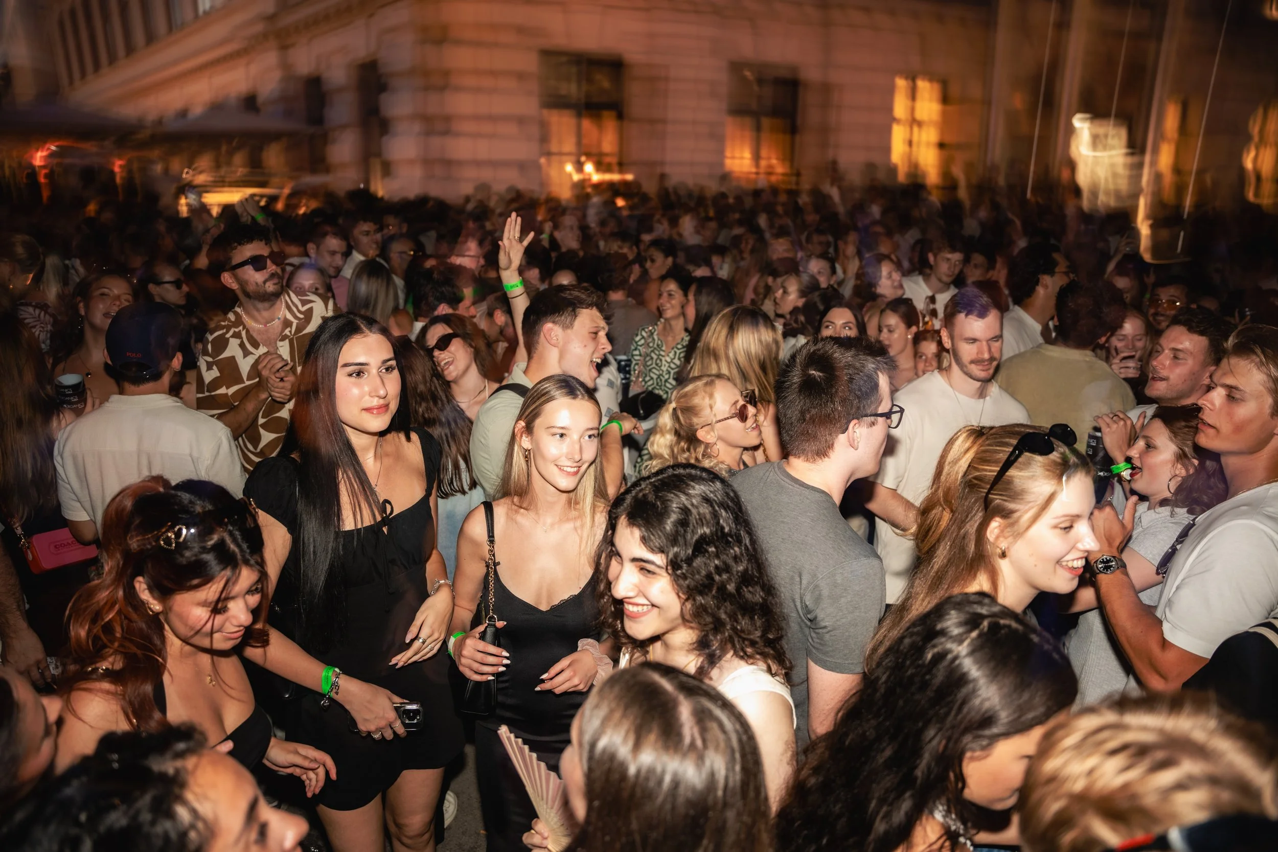 A large crowd of diverse young people enjoying a party or concert at night, with some wearing sunglasses and green wristbands.