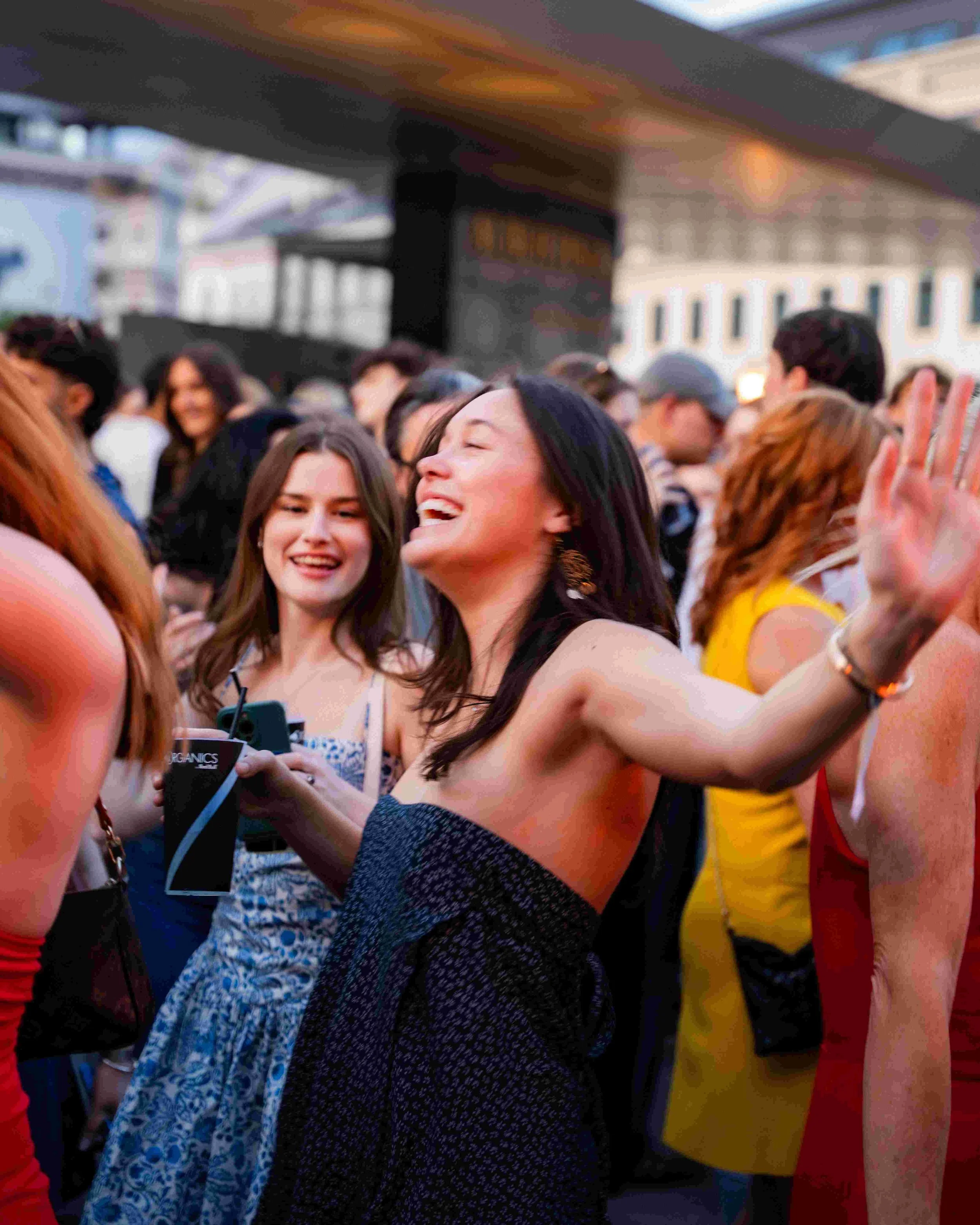 A group of people at an outdoor event, some smiling and taking selfies, with buildings and a stage in the background.
