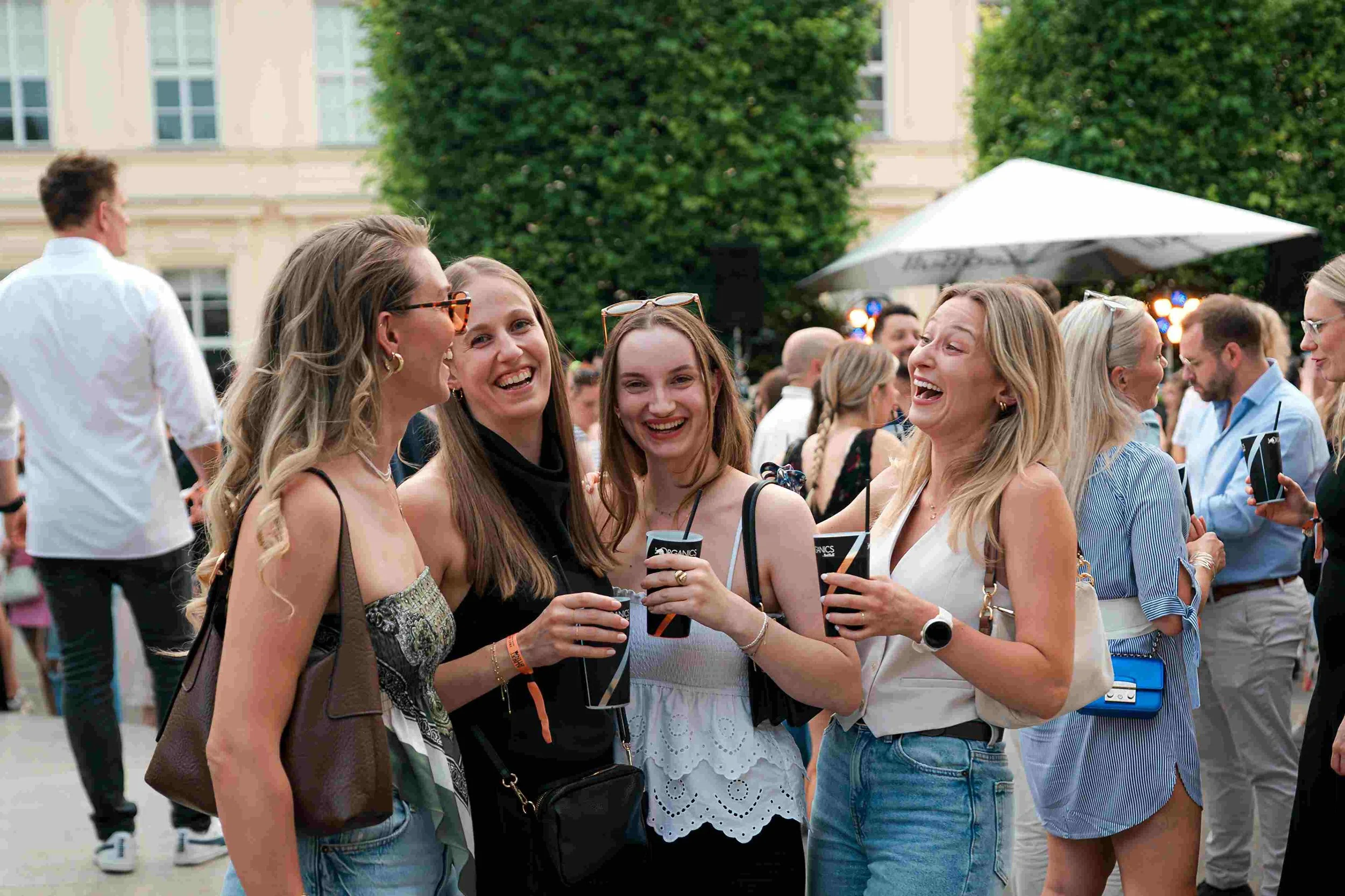 Group of smiling women talking and holding drinks at an outdoor event.