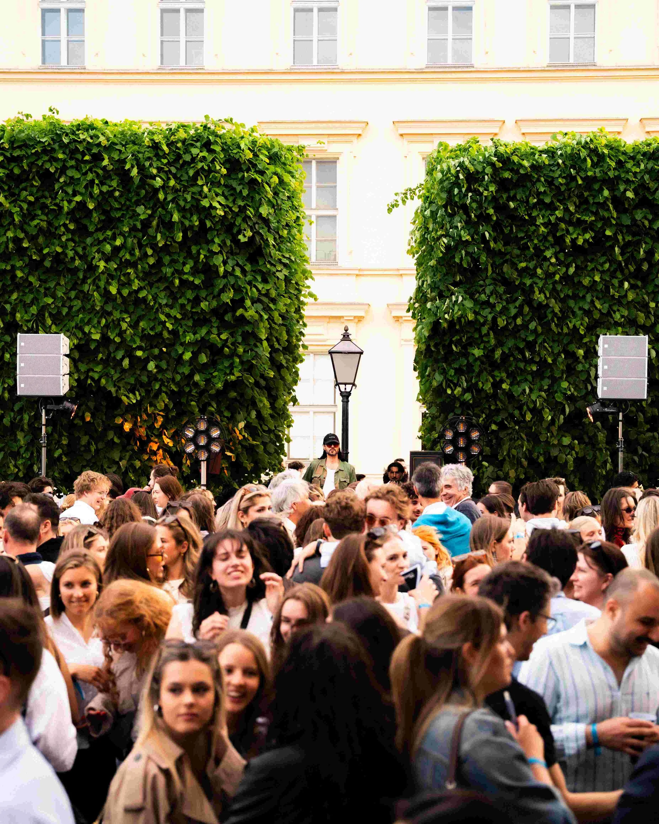 Crowd of people gathered outdoors in front of a building with large windows and tall hedge plants, with stage lighting and speakers set up, possibly for an event or performance.