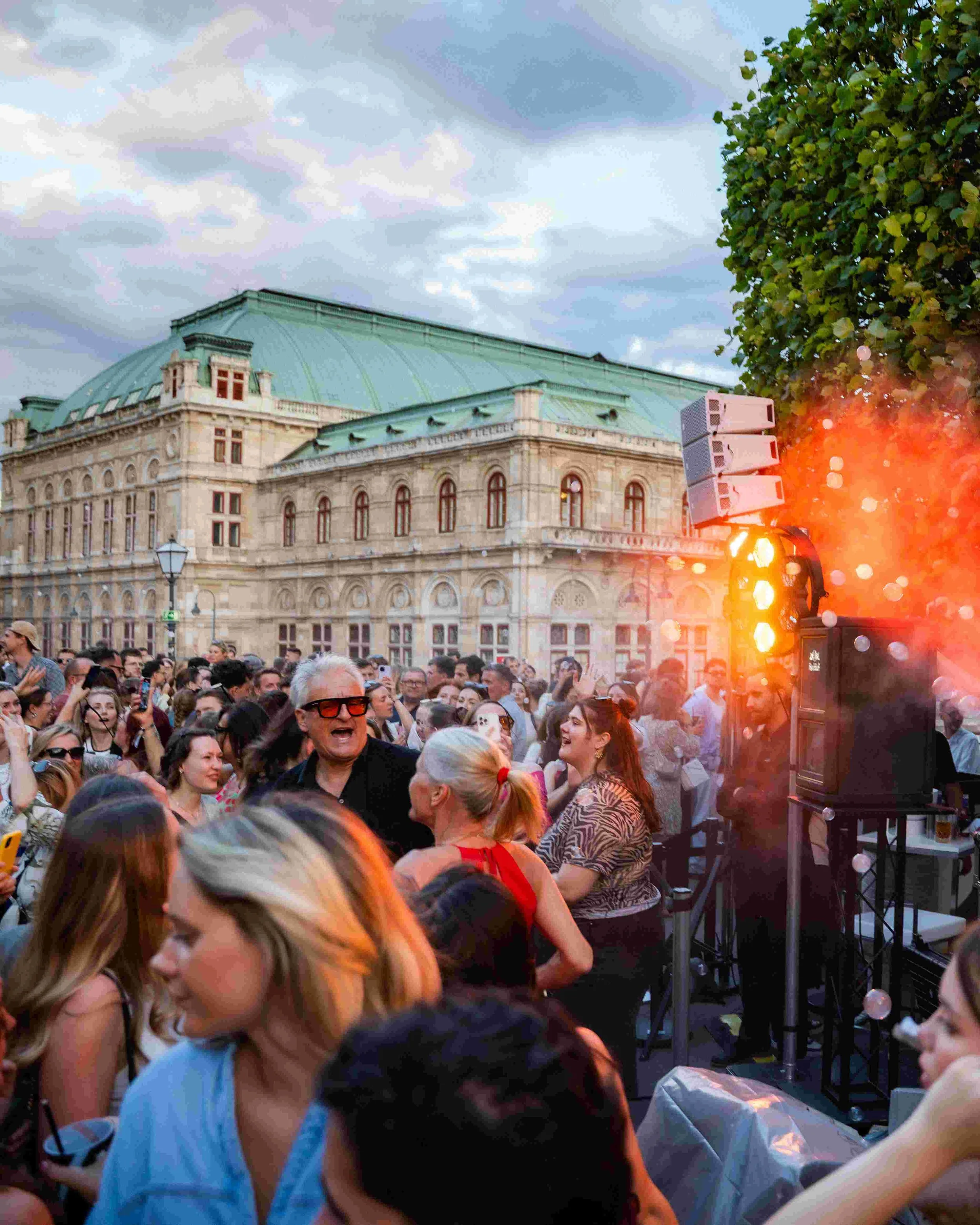 A crowd of people gathering outdoors during the evening, with a historic building featuring a green roof and ornate window arches in the background. The scene includes stage lighting and a large speaker setup on the right, with some individuals takin
